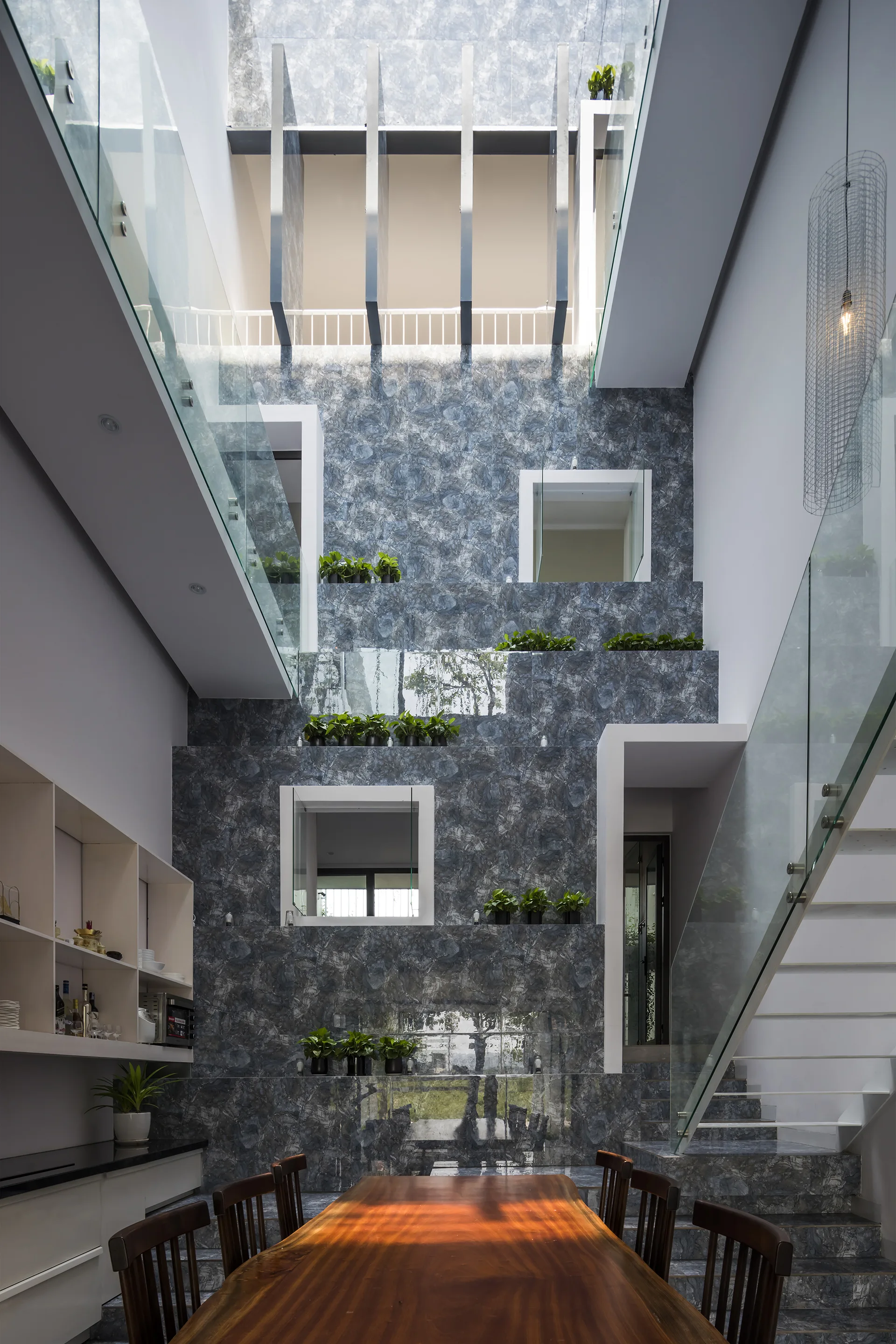 A modern dining area features a wooden table and chairs inside a tall atrium with blue-grey marble-patterned walls, glass railings, and square windows.
