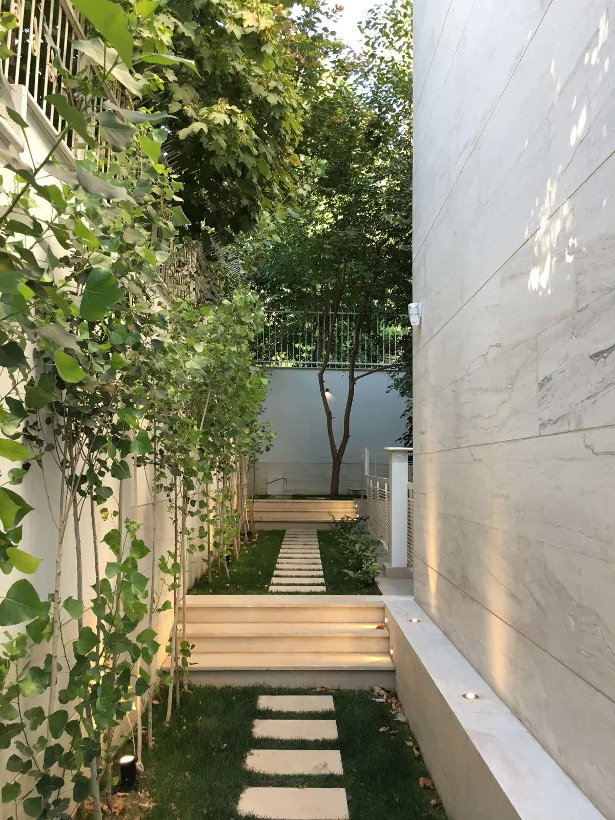 A narrow outdoor garden path with rectangular stone pavers in the grass, transitioning up stone steps alongside a tall, light-toned stone wall.