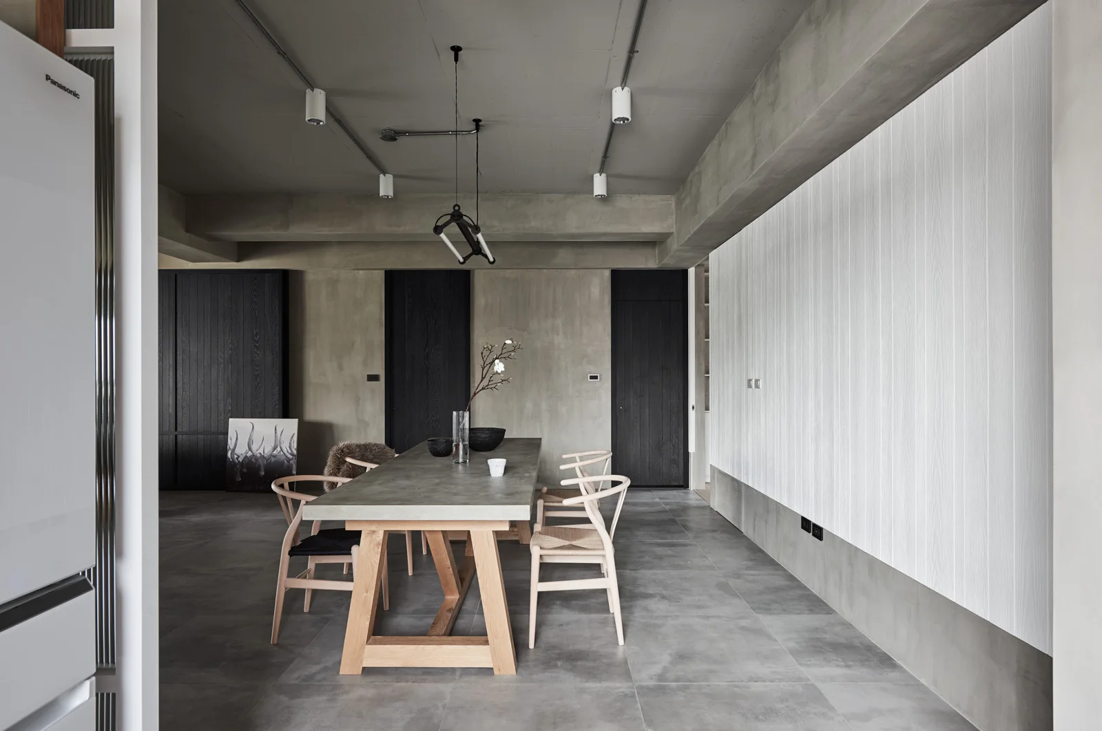 A modern dining room features an oak trestle table with light wood chairs under an exposed concrete ceiling with industrial track lighting.