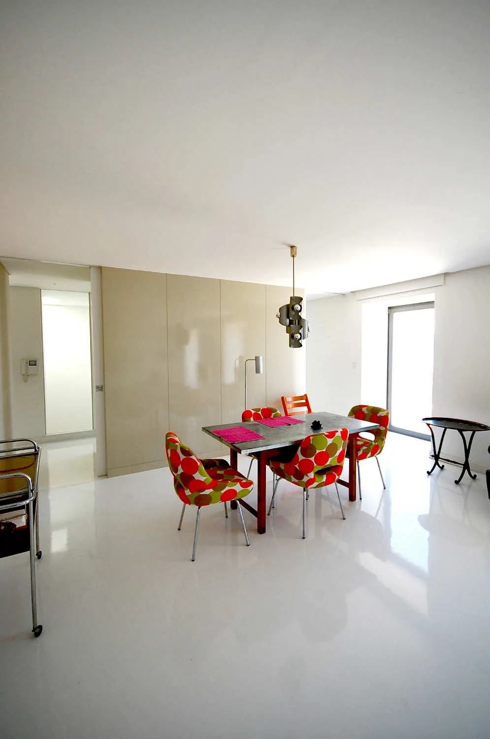 A minimalist dining room features a metal-topped table surrounded by four colorful polka-dot chairs on a glossy, highly reflective white floor.