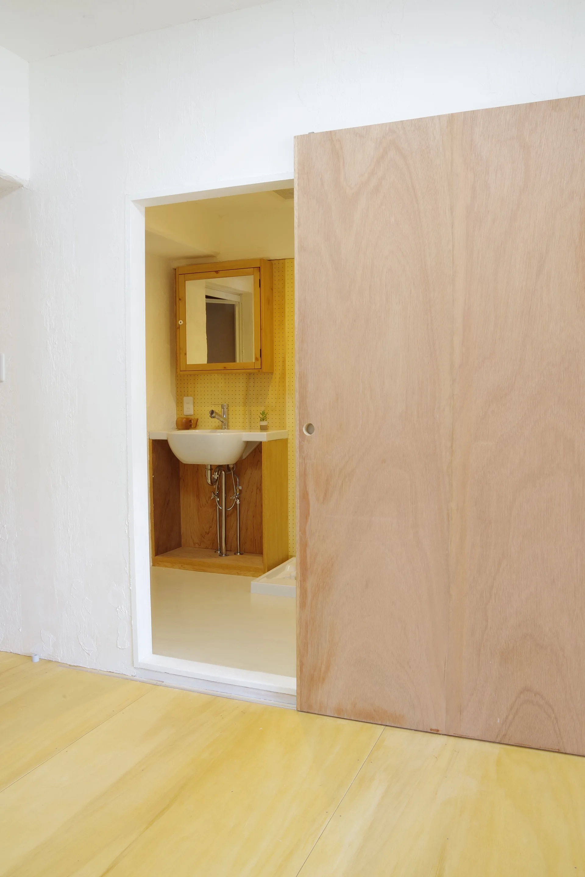 A view through a doorway into a small bathroom with a yellow pegboard wall, a wood vanity, and a large sliding plywood door.