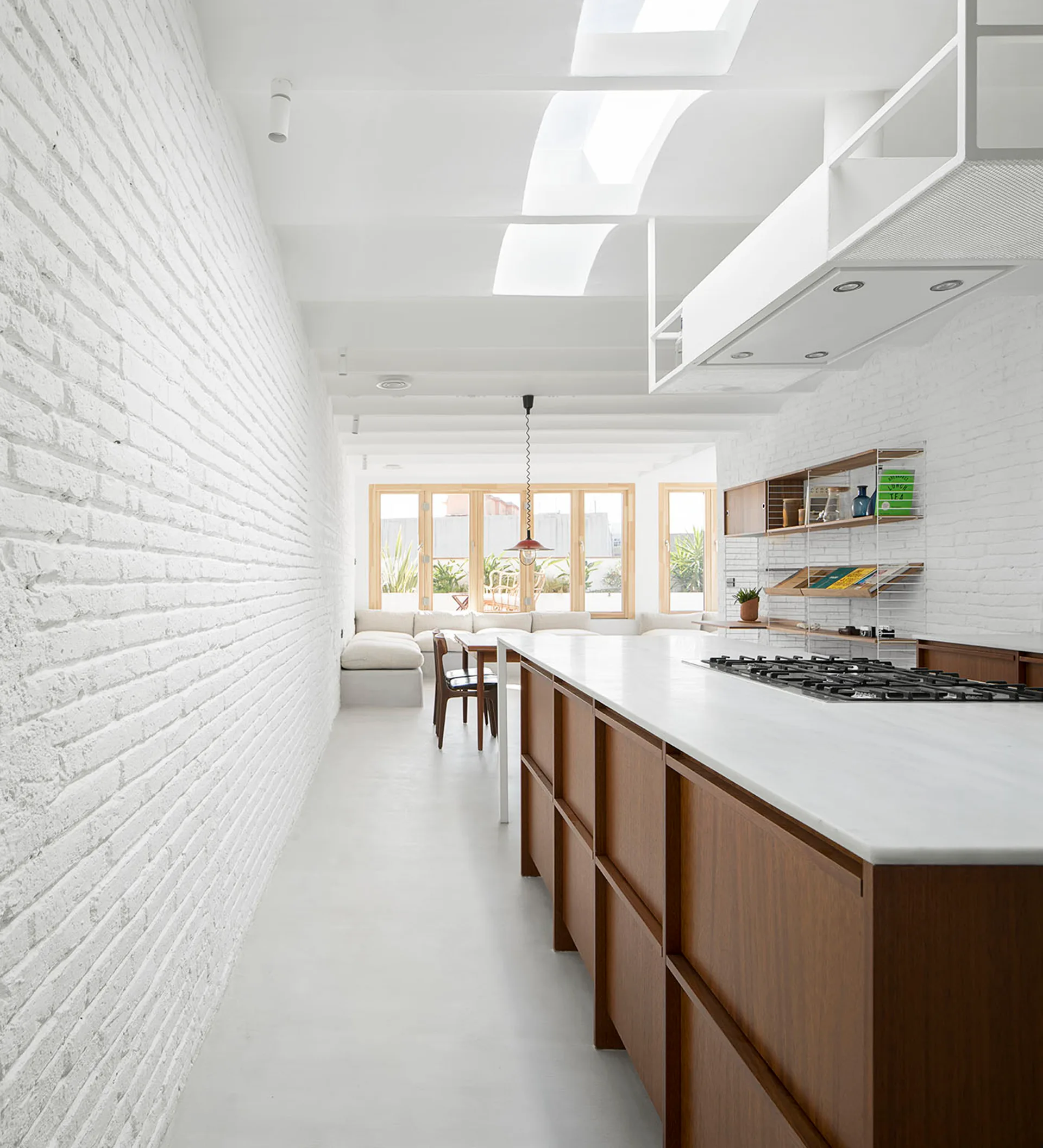 A minimalist kitchen featuring a long wood-toned island with a white top, textured white brick walls, ceiling skylights, and a seating area in the distance.
