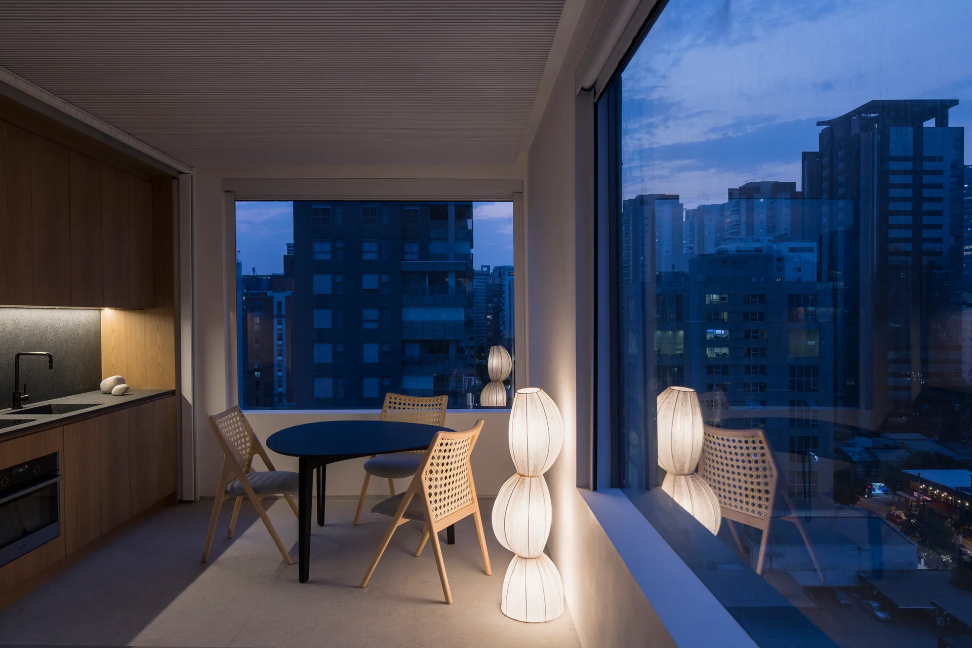 A contemporary dining area features a round black table, woven cane chairs, a sculptural floor lamp, and large windows overlooking a city skyline at dusk.