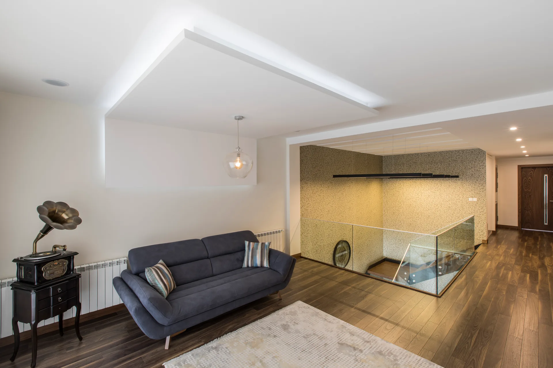 A dark grey sofa sits on wood floors next to a glass-railed staircase landing under a dropped ceiling panel with glowing cove lighting.