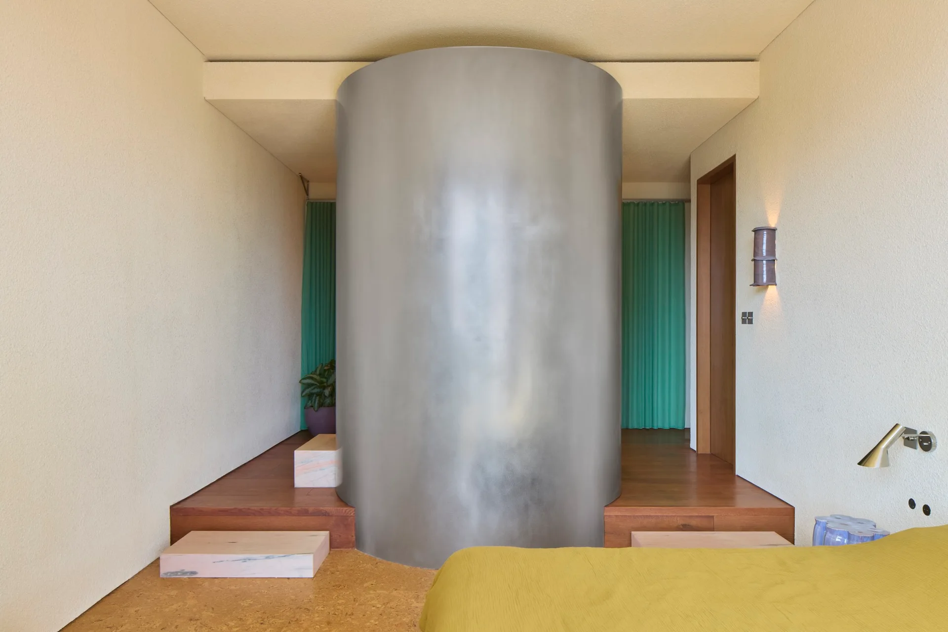 A modern bedroom features a large brushed metal cylinder, a yellow bed, a raised wooden platform, and green curtains in the background.