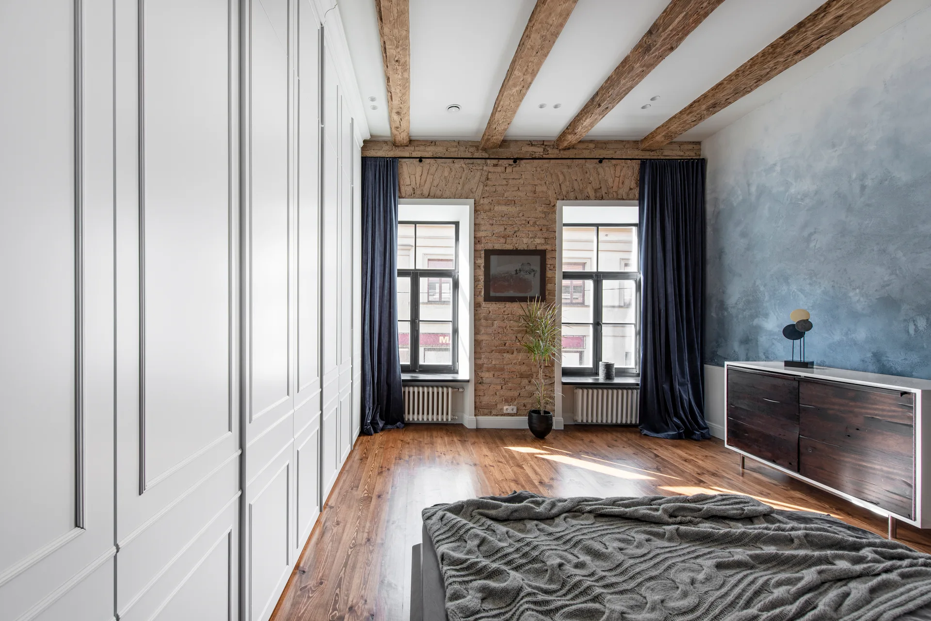 A modern industrial bedroom featuring an exposed brick wall, dark wood ceiling beams, a large white paneled wardrobe, and a blue ombre accent wall.