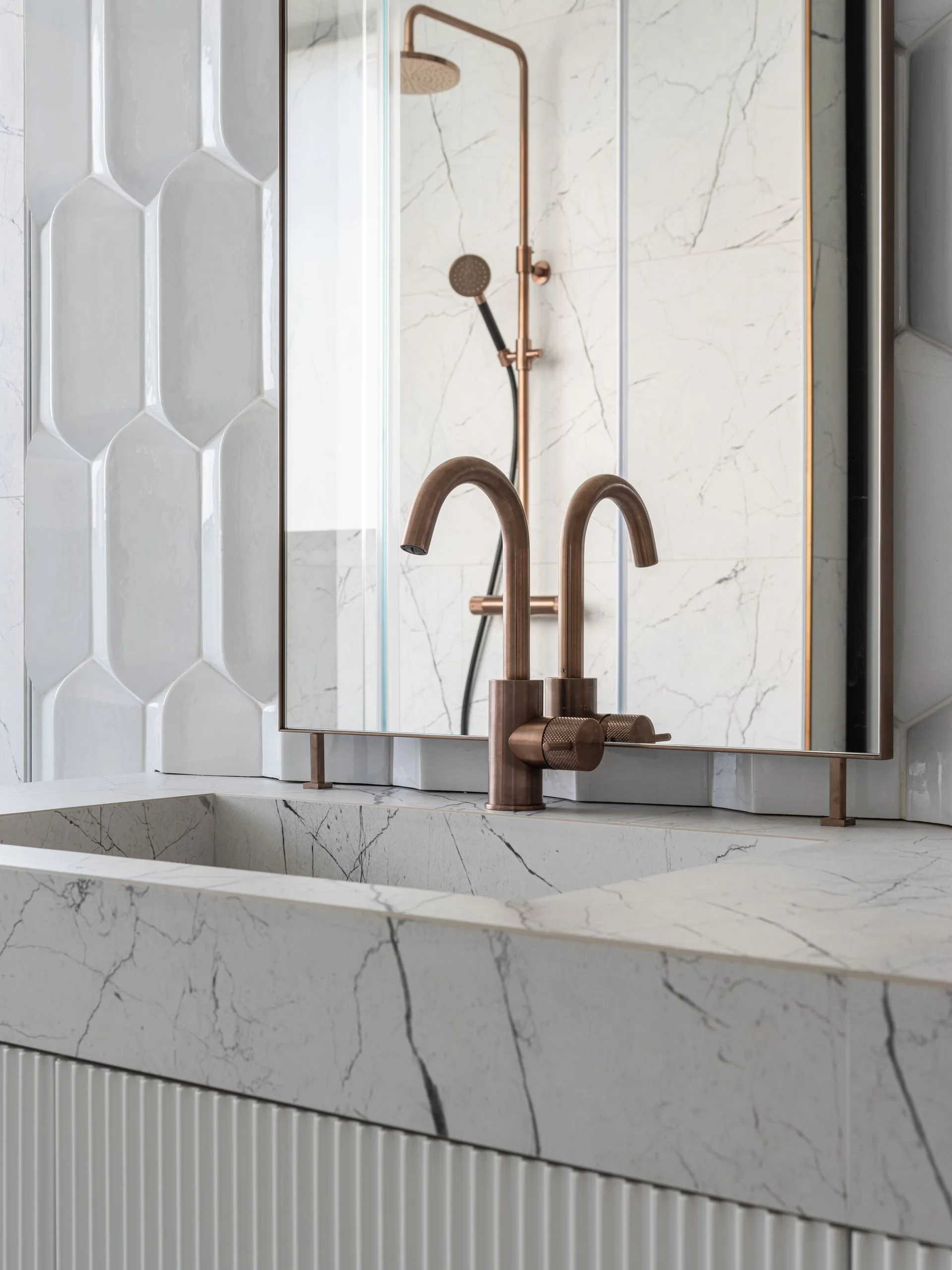 Close-up of a white veined marble bathroom vanity with an integrated sink, brushed copper faucet, rectangular mirror, and textured white wall tiles.