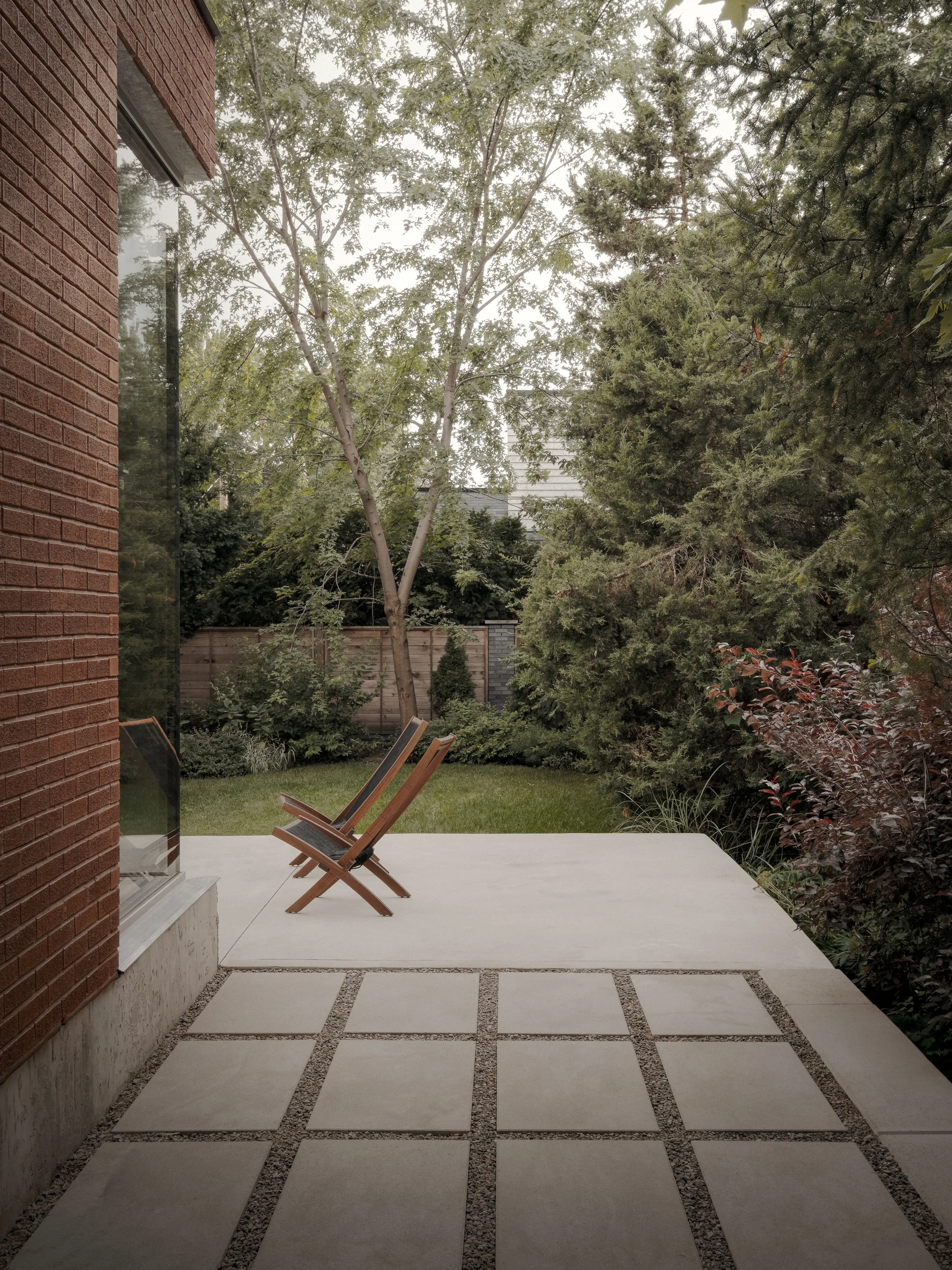 A grey concrete paver patio with wooden folding chairs sits next to a red brick house wall, facing a lush backyard with mature green trees.