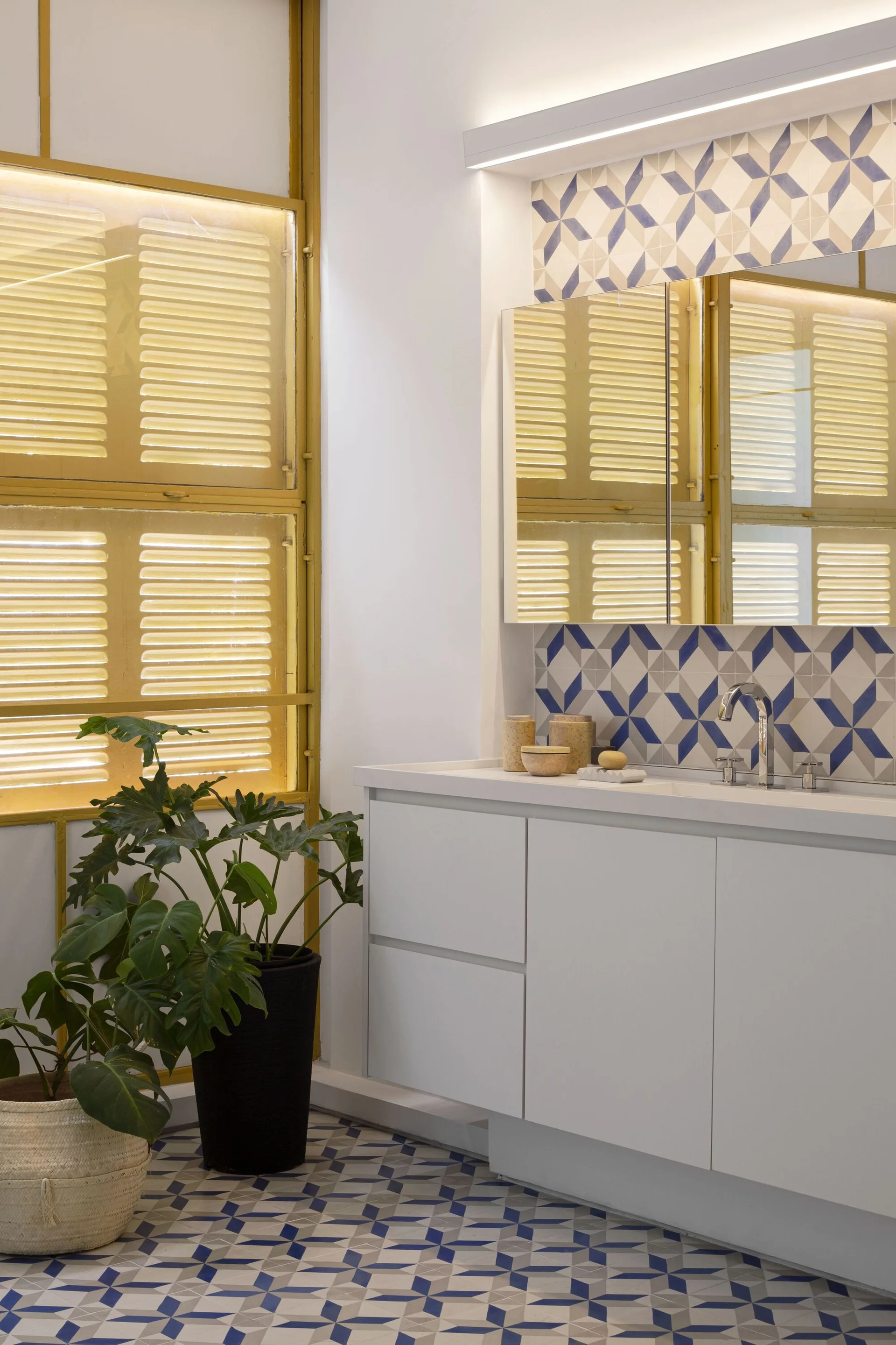 A modern bathroom vanity with a white cabinet and a blue and white geometric tile backsplash, next to a large window with yellow shutters.