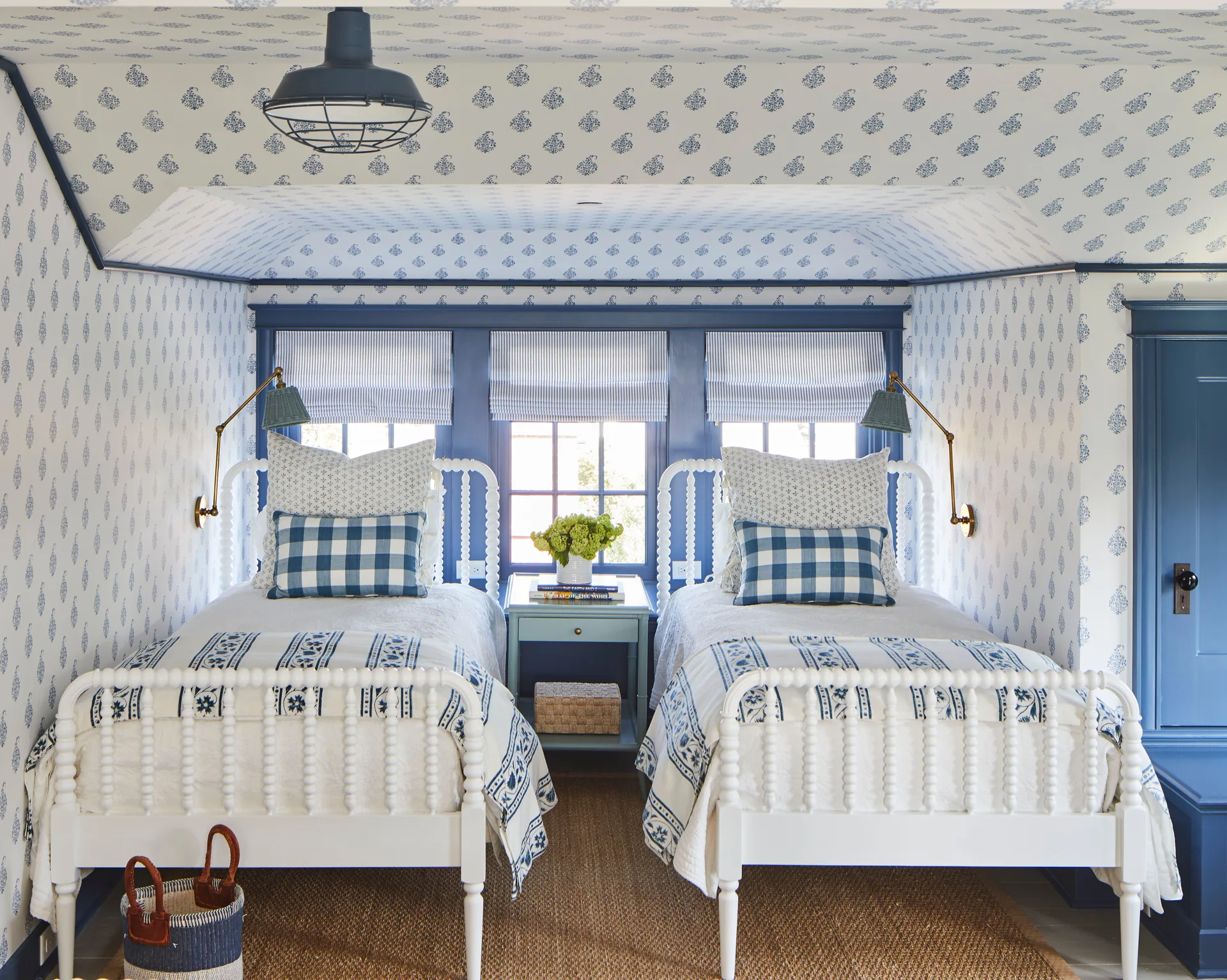 Two white spool beds stand symmetrically in an attic bedroom covered in blue and white floral wallpaper, framing a blue-trimmed window niche.