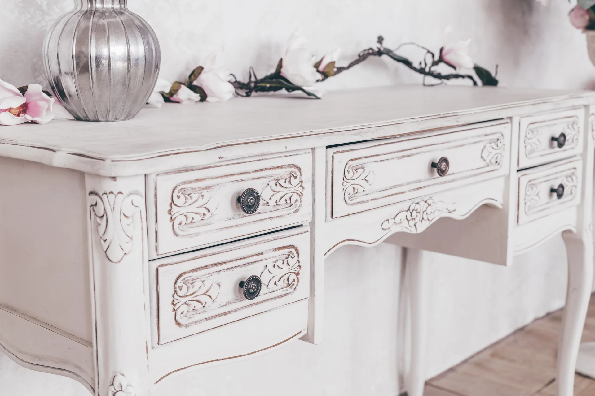 A distressed white wood console table with ornate carved drawers, a silver metallic vase, and faux pink magnolia blossoms on top.