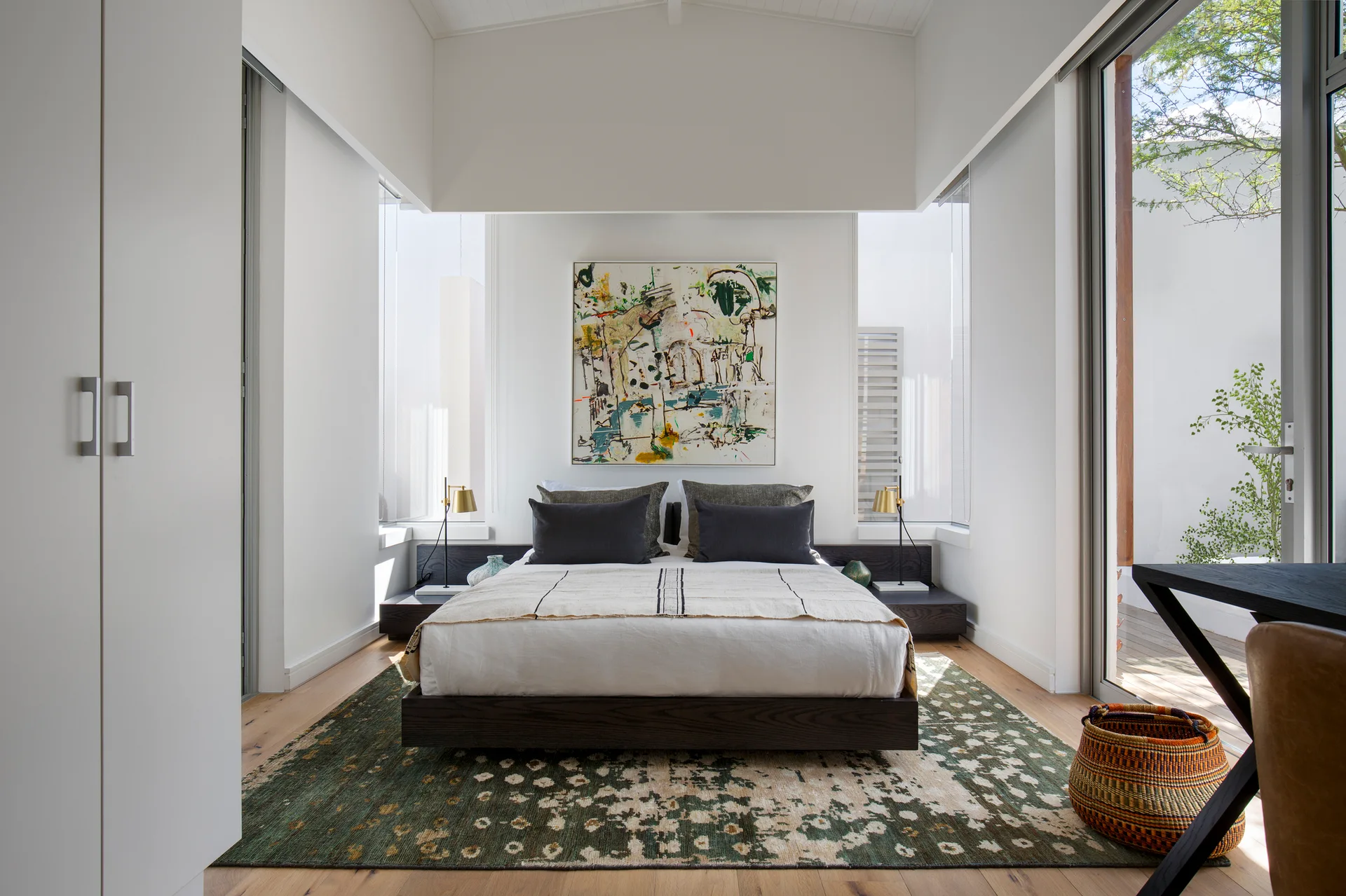 A modern bedroom featuring a low dark wood bed frame centered between two tall narrow windows with abstract art on the wall above.