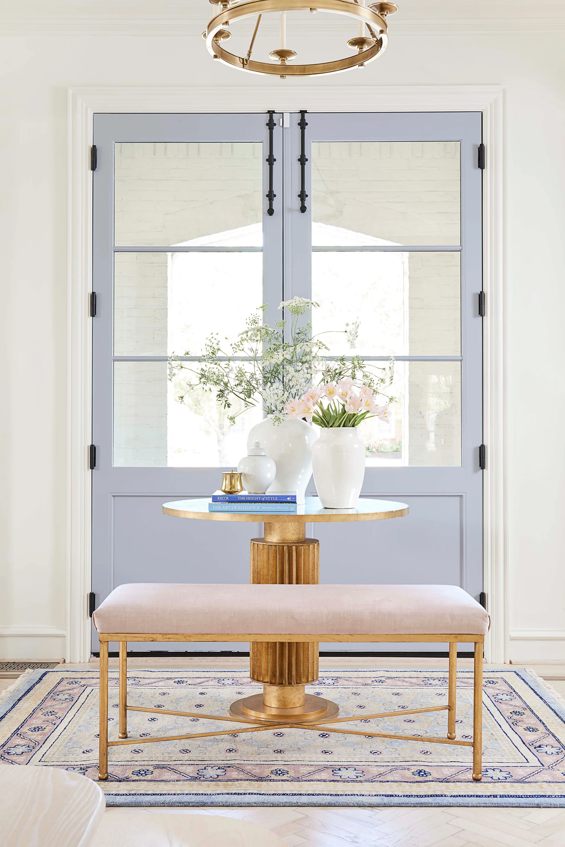 A light pink upholstered bench and a gold pedestal table with white vases sit in front of light blue double doors in an entryway.