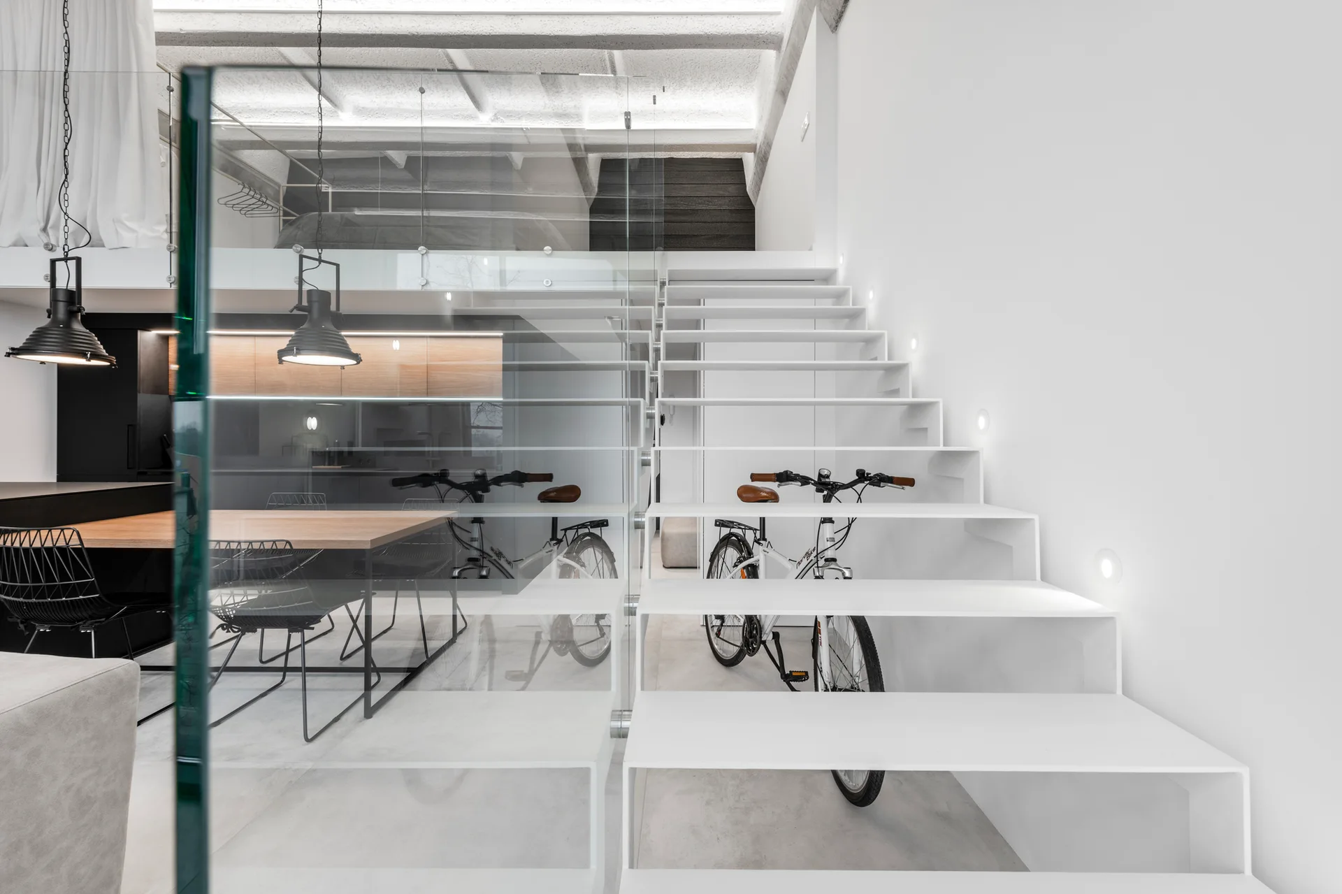 A minimalist white metal floating staircase with a transparent glass railing and recessed wall lights in a modern loft.