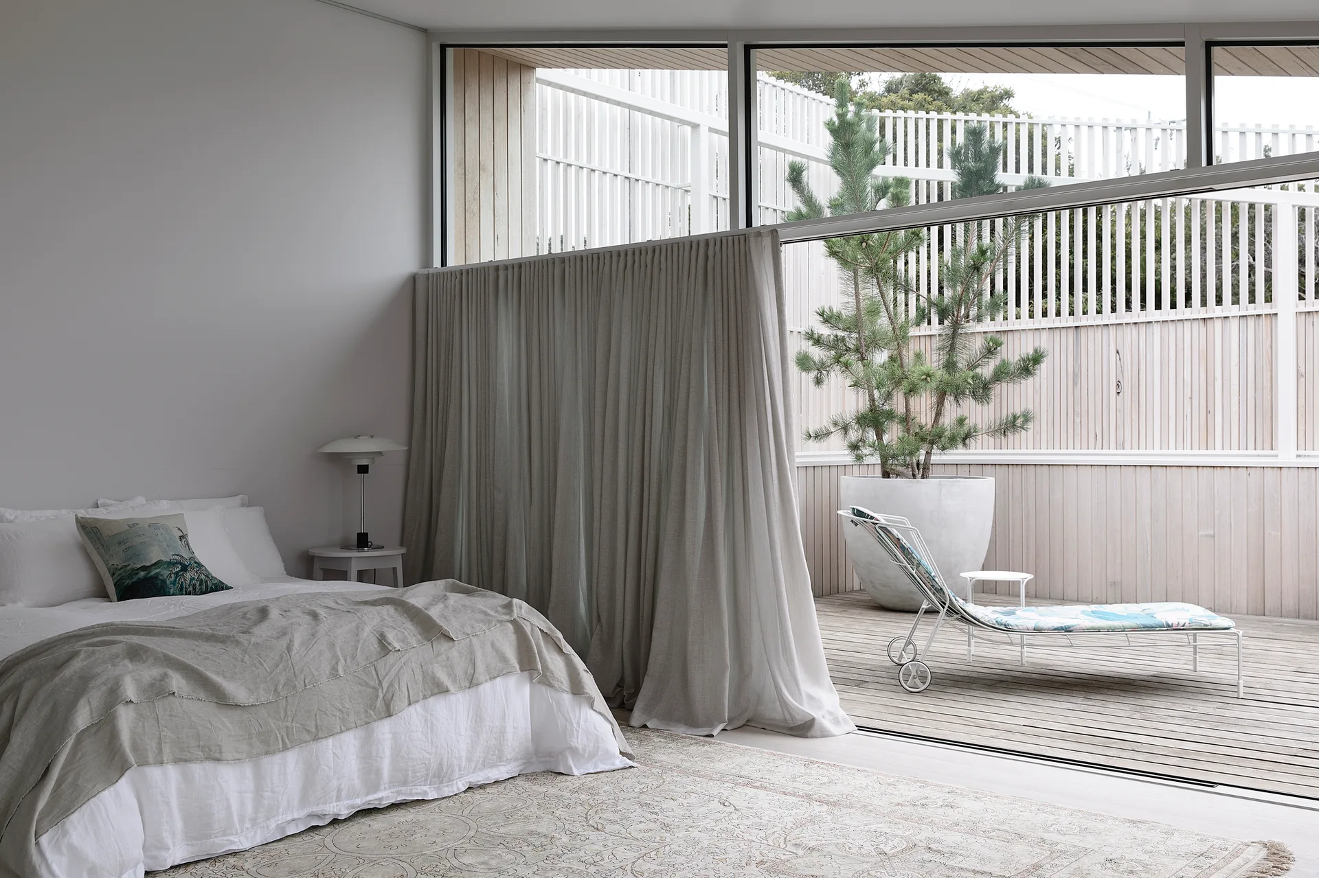 A minimalist bedroom with white linen bedding opens through sliding glass doors to a wooden deck with a potted conifer and lounge chair.