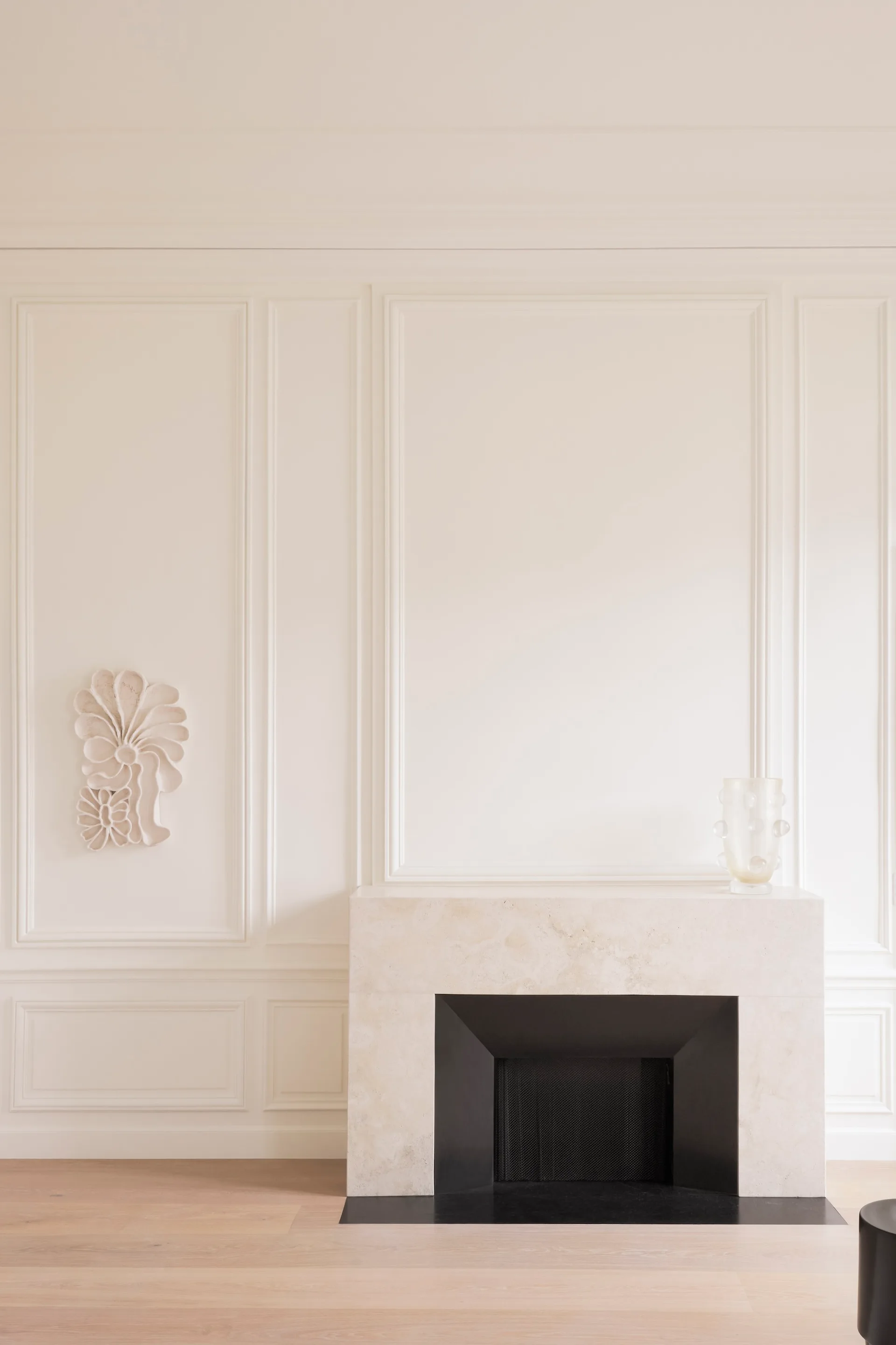 A minimalist cream stone fireplace is centered below white paneled walls adorned with a small plaster floral sculpture and a textured glass vase.