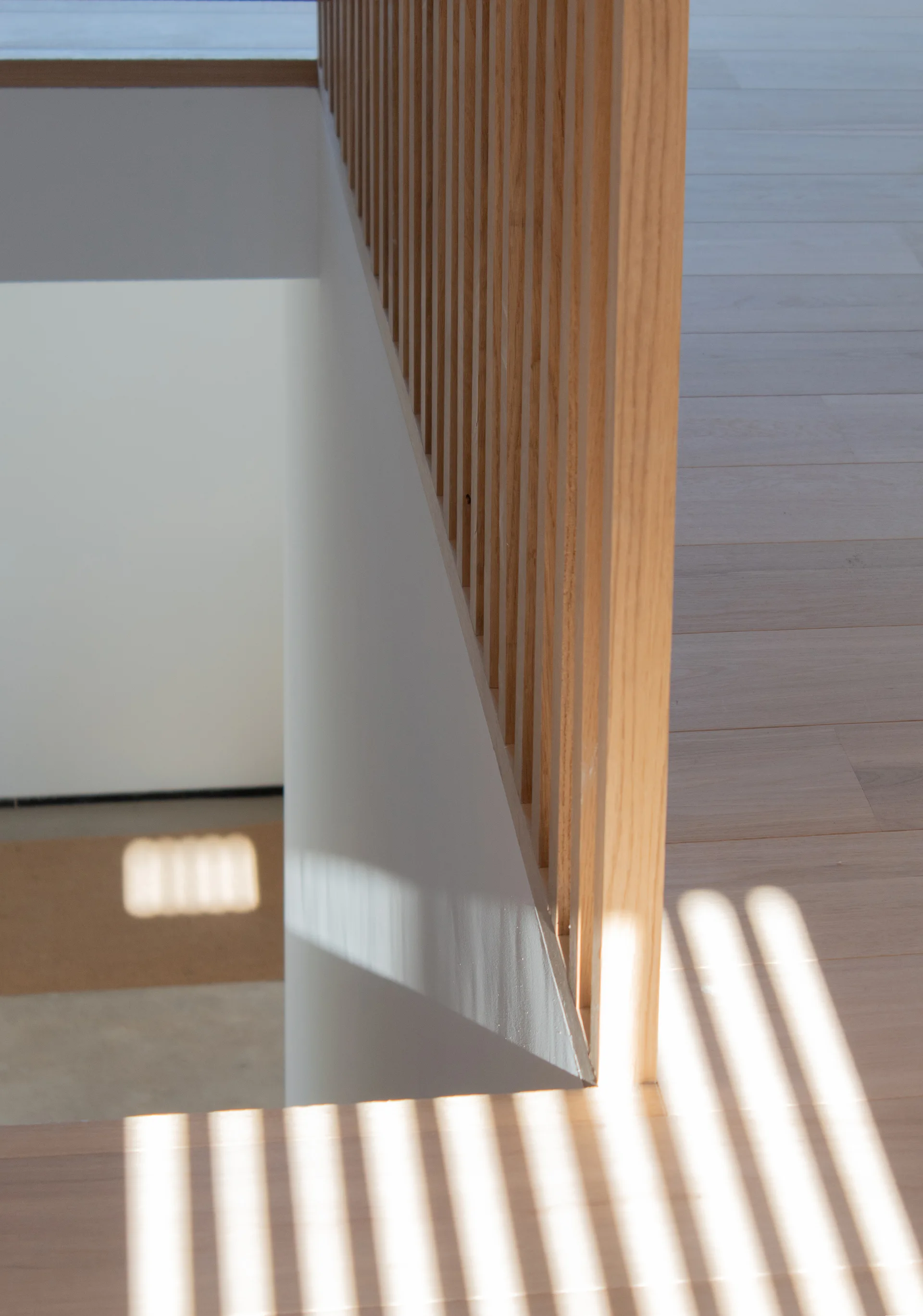 A minimalist staircase features a vertical light wood slat railing casting striped shadow patterns onto a white wall and wood plank flooring.