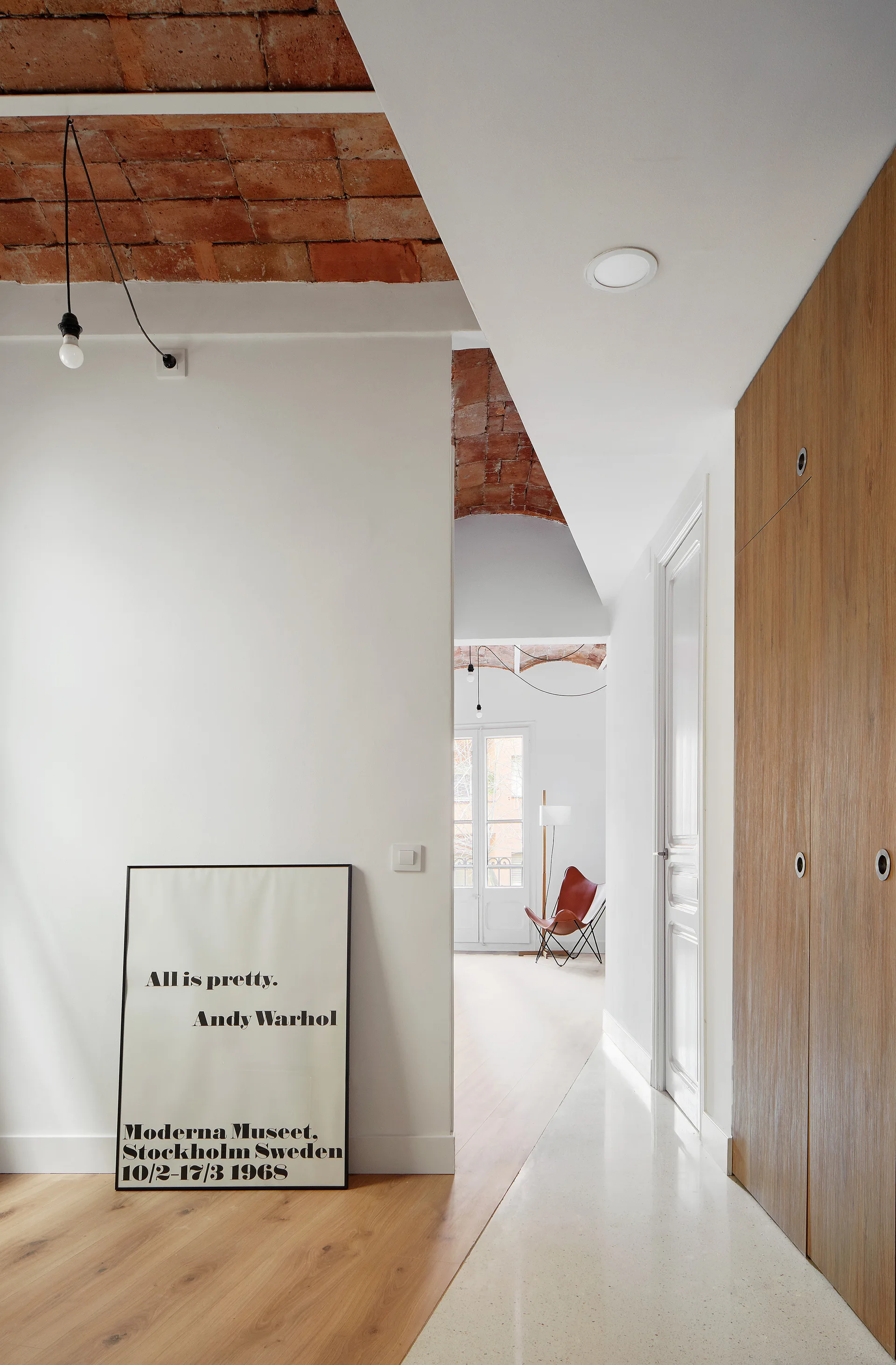 A bright interior hallway features a vaulted brick ceiling, flush-mount wooden cabinets, and a framed art poster leaning against a white wall.