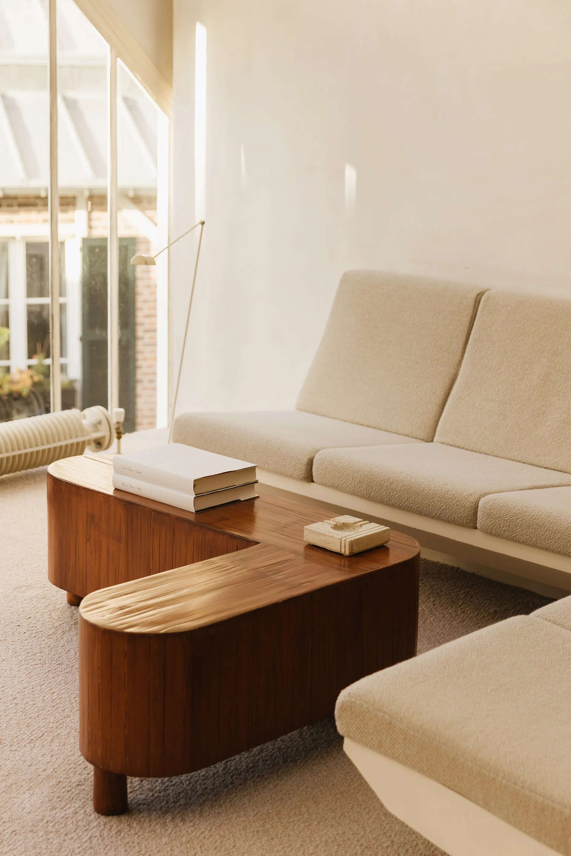 A low-profile textured cream sofa sits beside a curved wood coffee table in a sunlit room with an arched window.