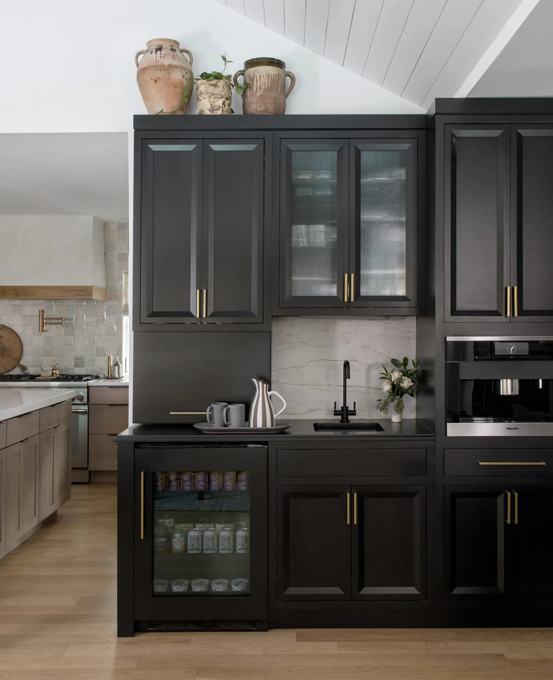 Custom black cabinetry surrounds a built-in coffee machine, bar sink, glass-door beverage fridge, and marble backsplash under a vaulted ceiling.
