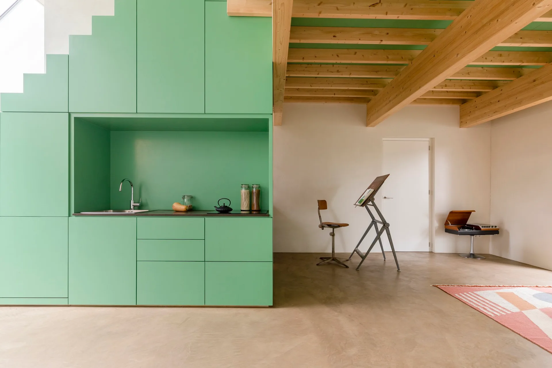 A minimalist open space features a bold green kitchen unit built under a staircase next to exposed wooden ceiling beams and a concrete floor.