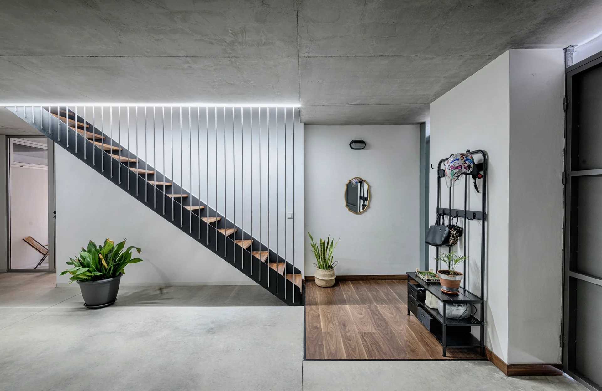 A modern entryway features a black floating staircase with vertical metal rod railings, concrete floors, a wood floor inlay, and a black coat rack.