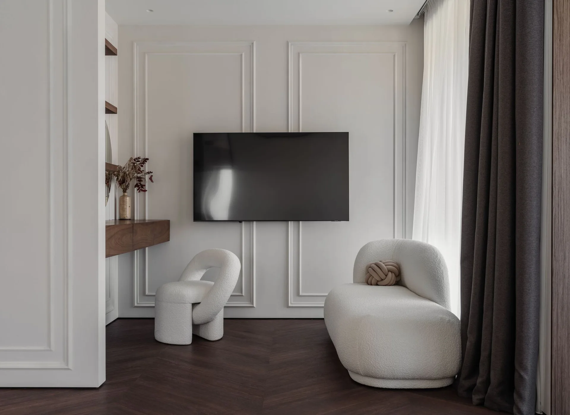 Sculptural white bouclé seating sits on dark wood herringbone flooring in a room with classic wall molding and a mounted television.