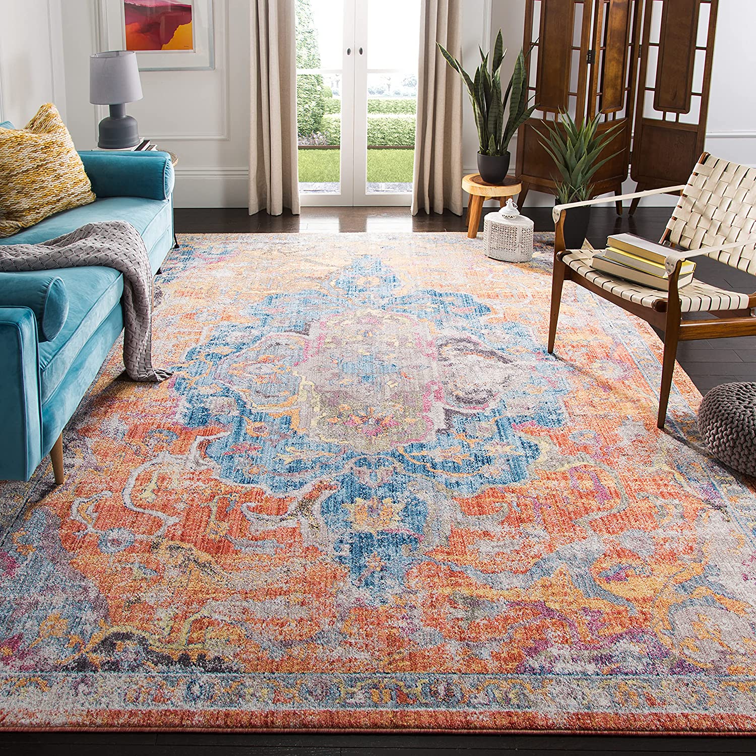 A living room featuring a colorful blue and orange medallion area rug, a teal sofa, a wooden accent chair, and double glass doors.