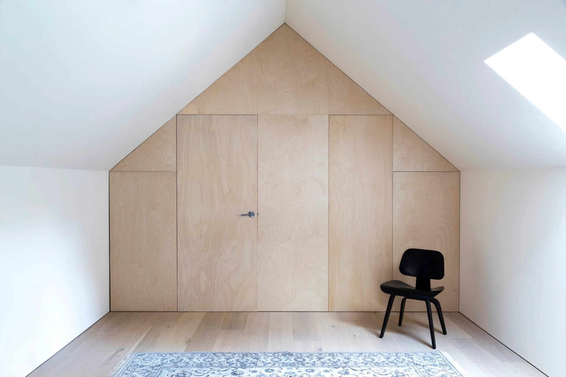 A minimalist attic bedroom featuring a light wood plywood paneled storage wall and a single black chair on a light wood floor.