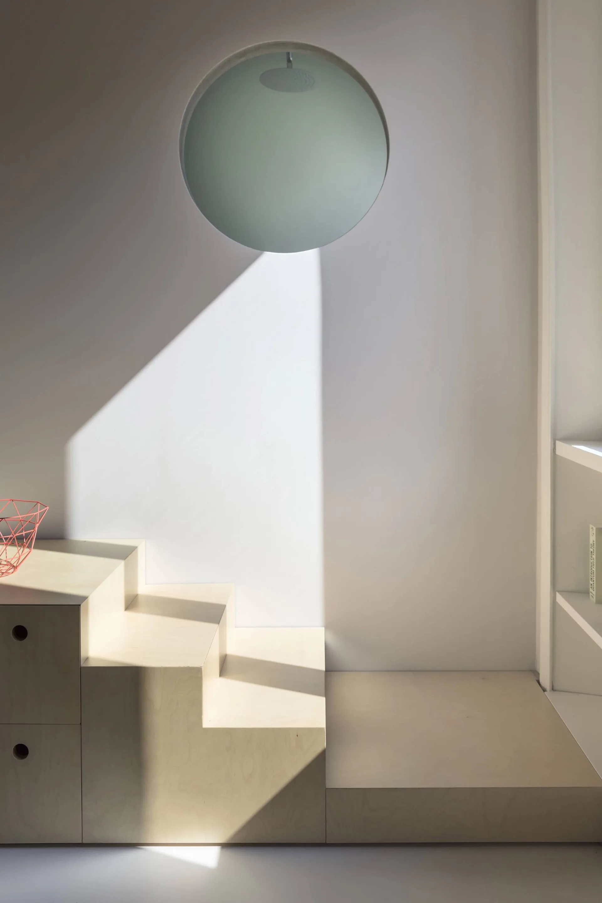 A stepped plywood storage unit with integrated drawers sits against a flat white wall beneath a circular interior window cutout.