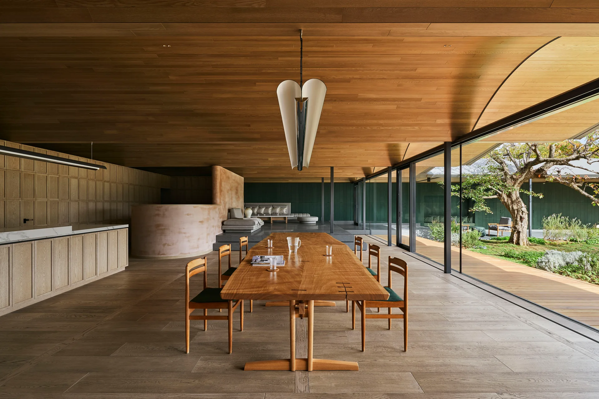 A long wooden dining table and chairs beneath a wood plank ceiling, with glass sliding doors opening to an outdoor courtyard.