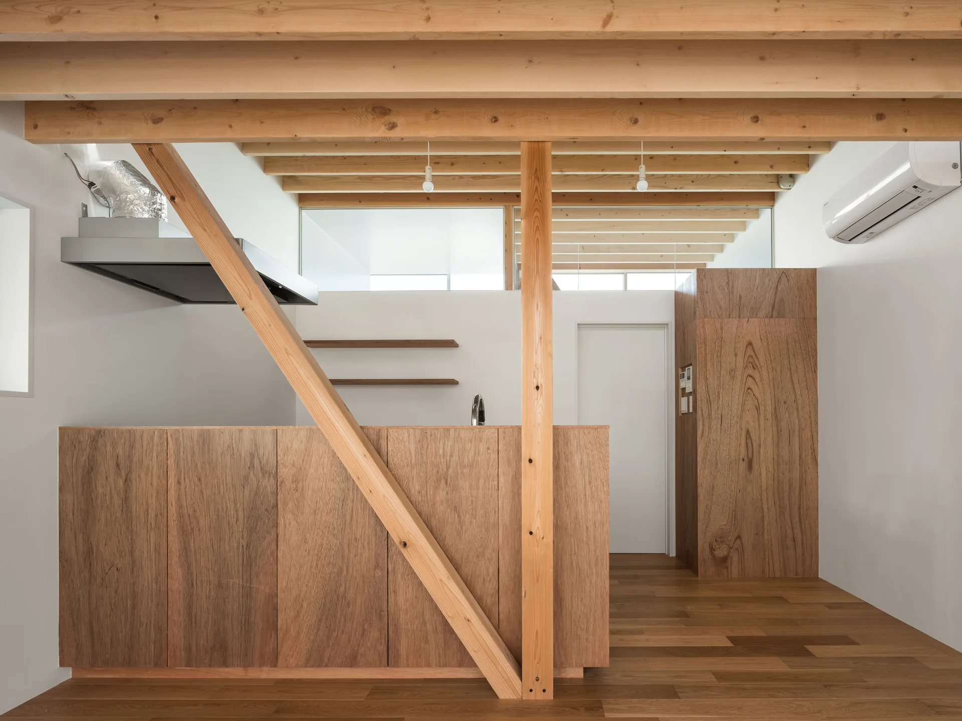 A minimalist kitchen features natural wood cabinets, exposed timber ceiling beams, and a diagonal support post against clean white walls.