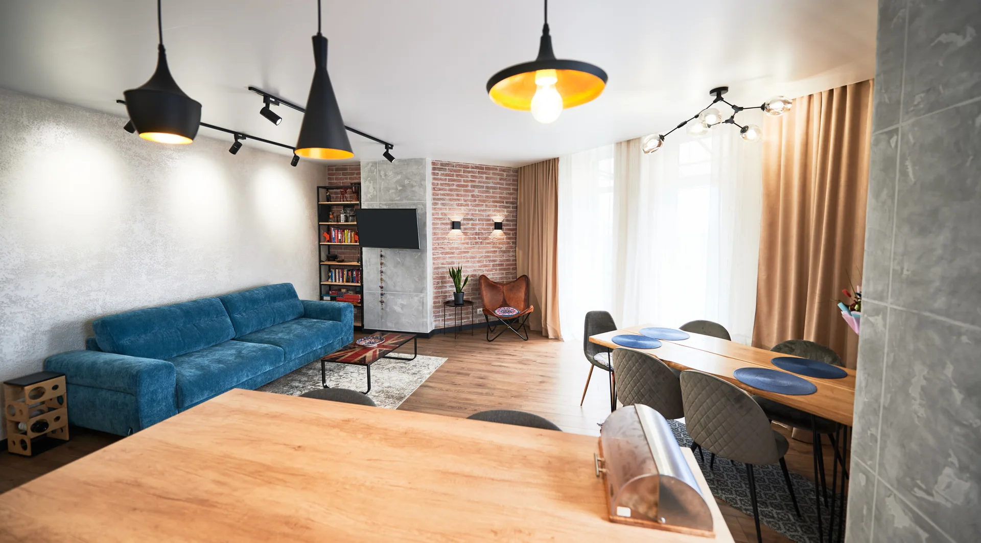 An open-plan residential interior features a blue fabric sofa, an exposed brick wall, black dome pendant lights, and a wooden dining table with grey chairs.