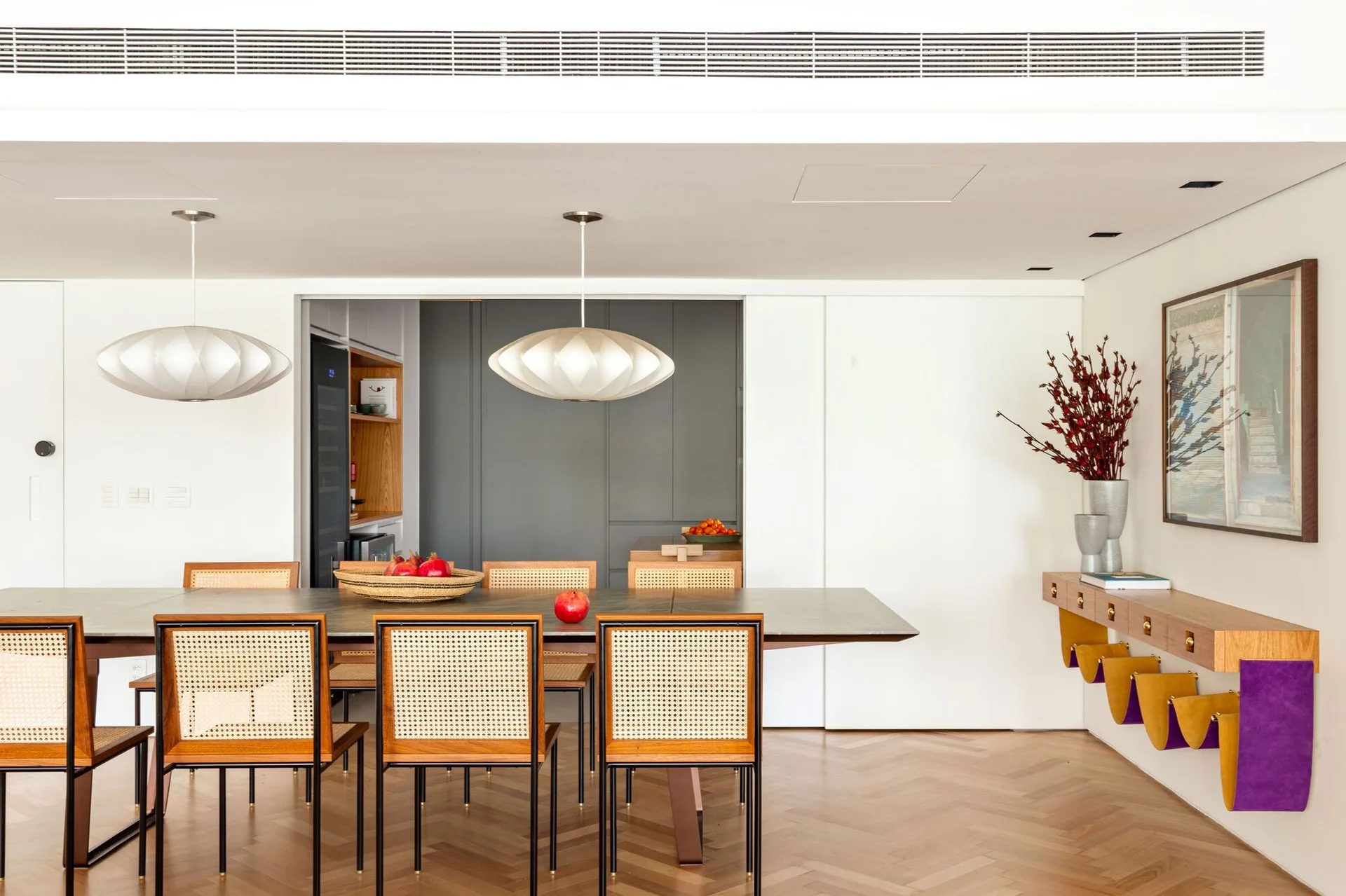 A modern dining room features a long table with wood and cane chairs under two pendant lights, alongside a floating wall shelf and herringbone flooring.