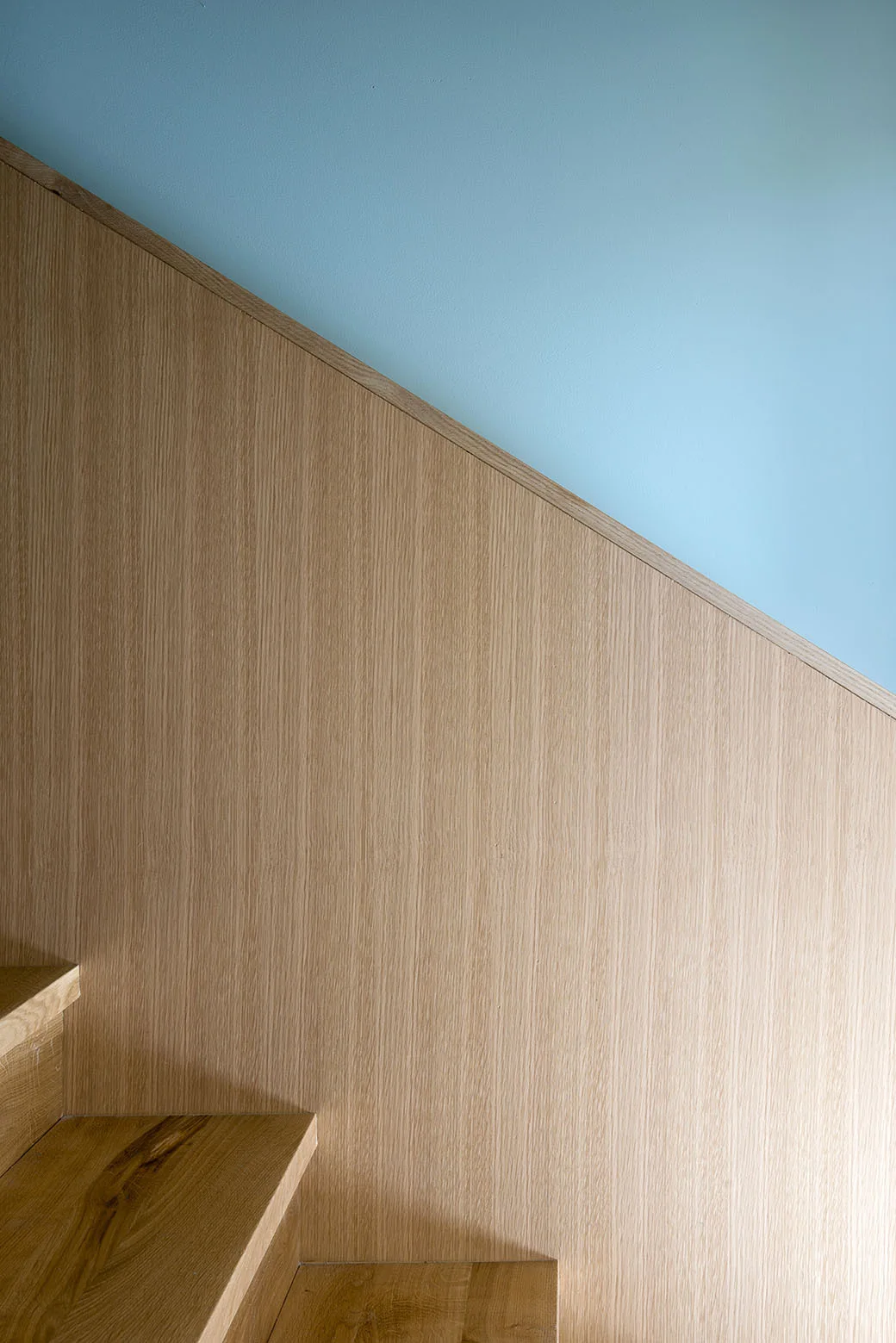 Wooden stair treads ascend beside a diagonal light oak wall panel that meets a soft blue painted surface.