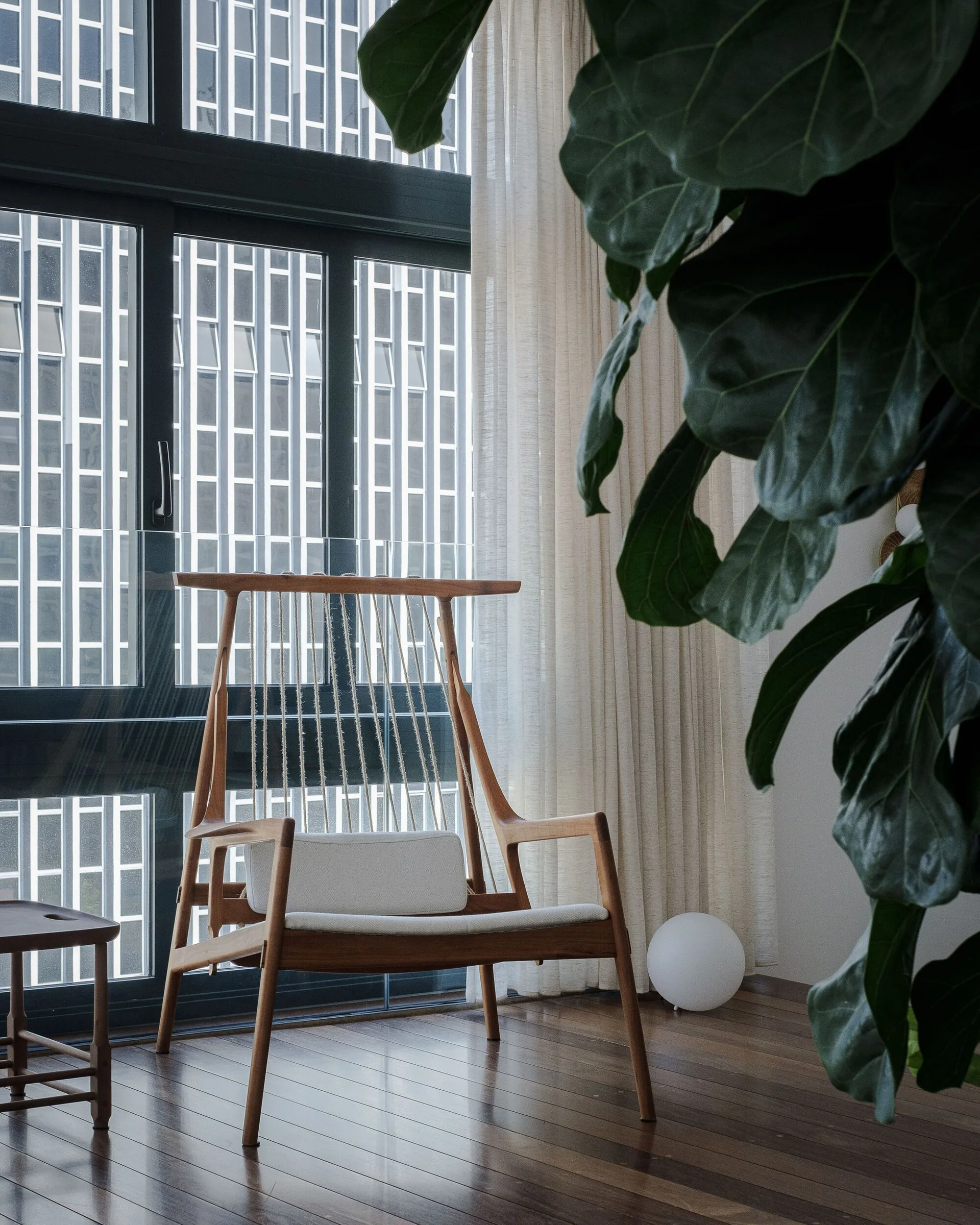 A wooden lounge chair with rope back detailing and a white seat cushion sits next to a large black-framed window.