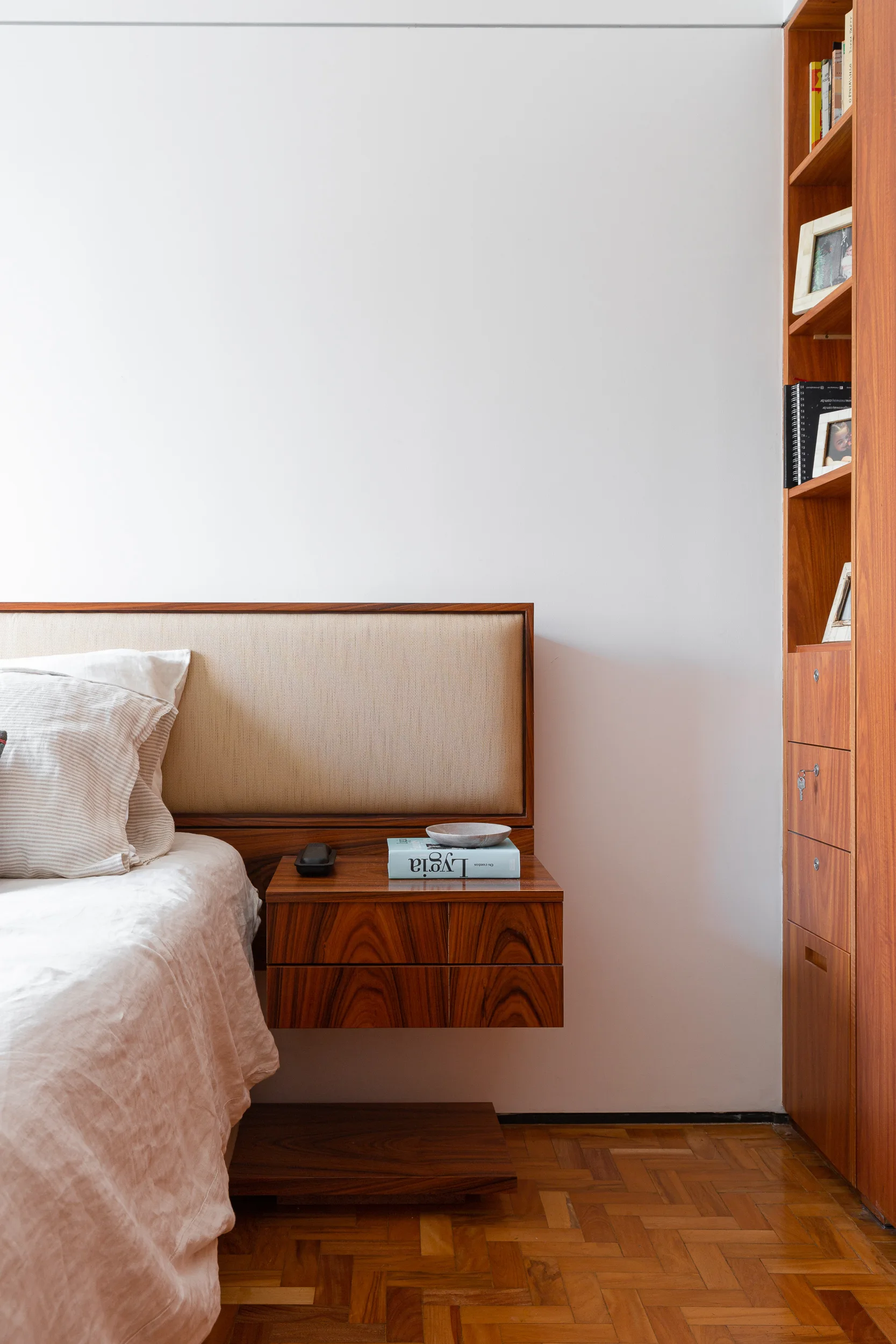 A minimalist bedroom corner features a floating wood nightstand integrated with a textured headboard, next to a tall wooden bookshelf and parquet flooring.