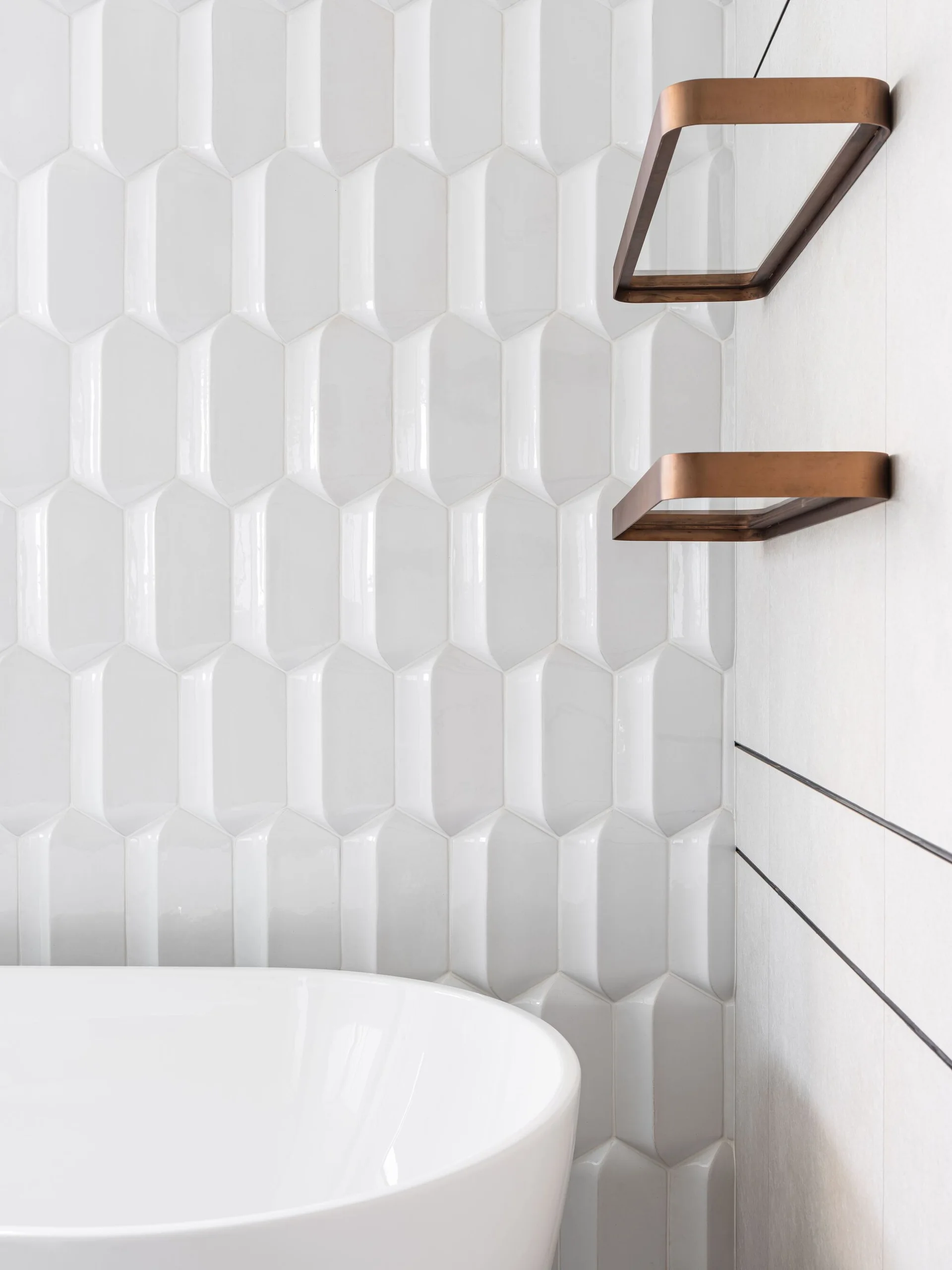 A bathroom corner featuring white glossy 3D geometric wall tiles, a portion of a white bathtub, and two copper-finished metal wall shelves.