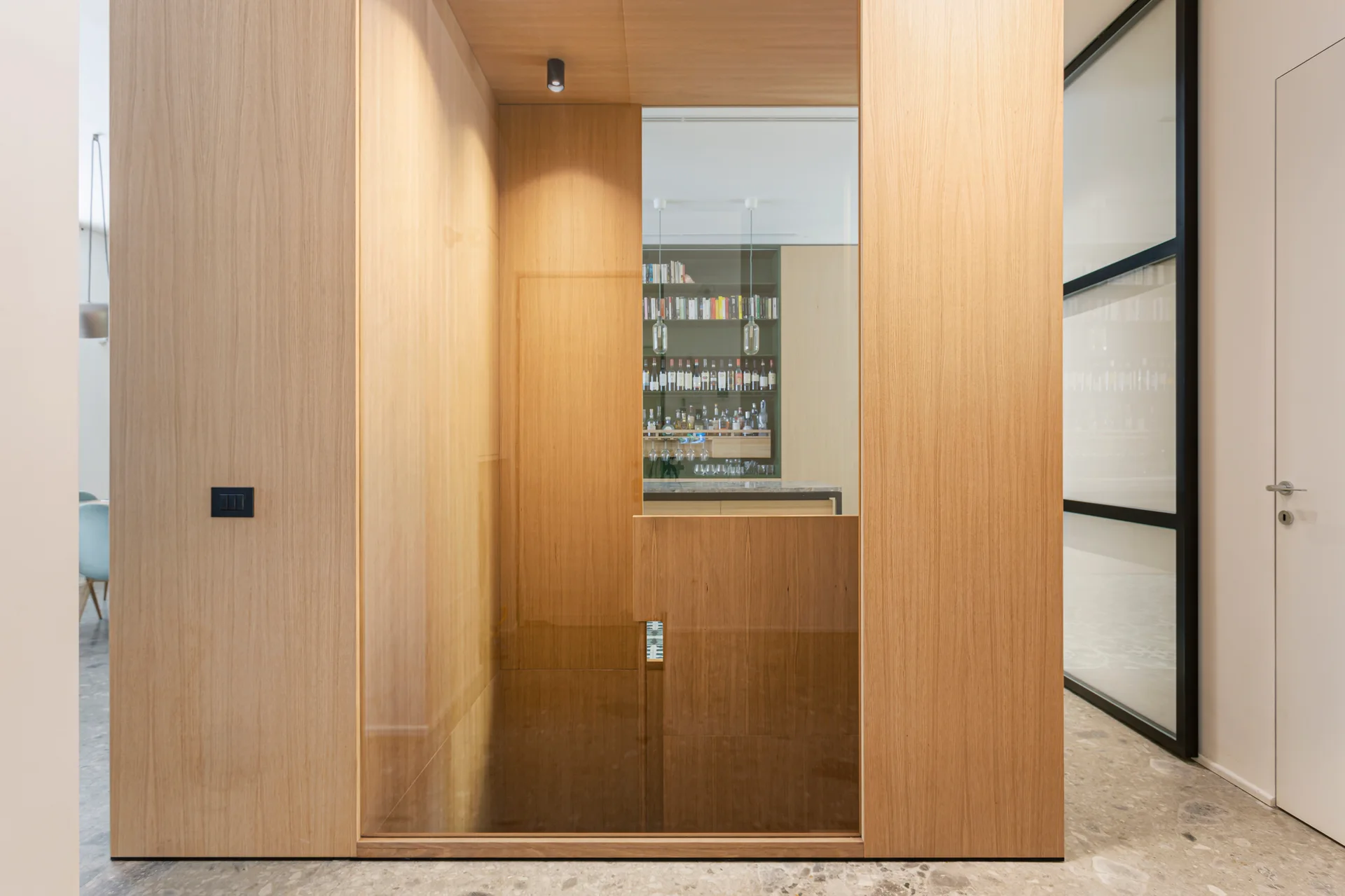 A floor-to-ceiling light wood partition wall features a clear glass window inset looking into a home bar with stocked shelves over speckled stone flooring.