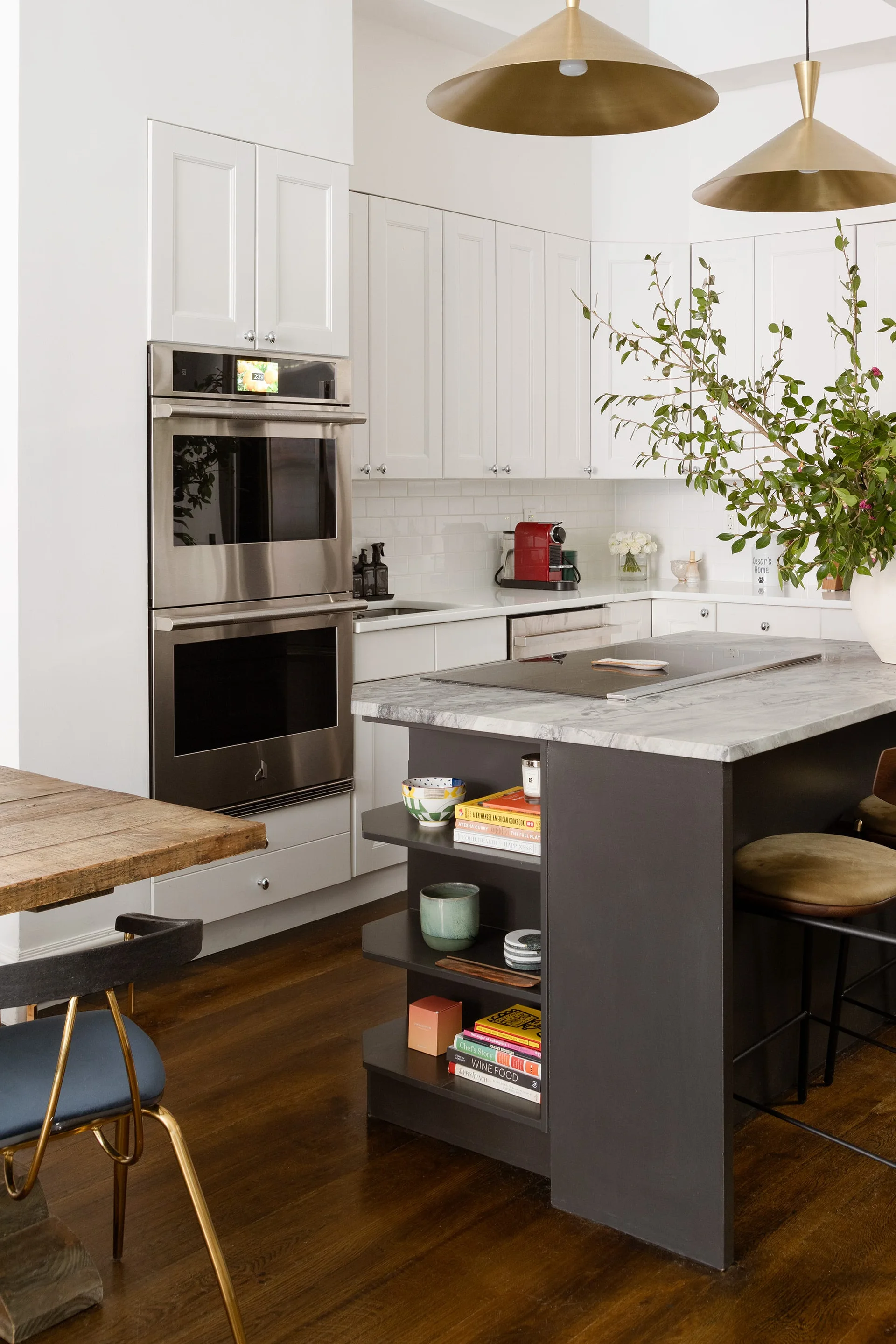 A modern kitchen features a dark gray island with open shelves, white shaker cabinets, stainless steel double ovens, and large brass pendant lights.