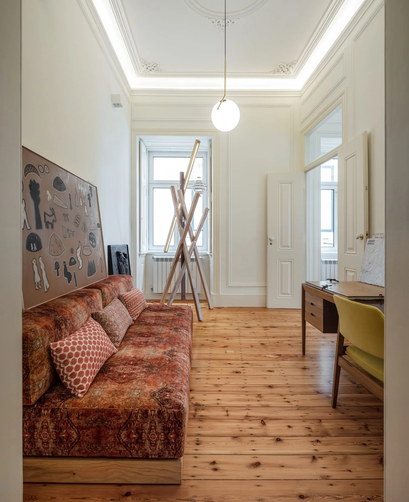 A bright living room features a long patterned rust-colored sofa, pine wood plank flooring, a small wooden desk with a yellow chair, and high coved ceilings.