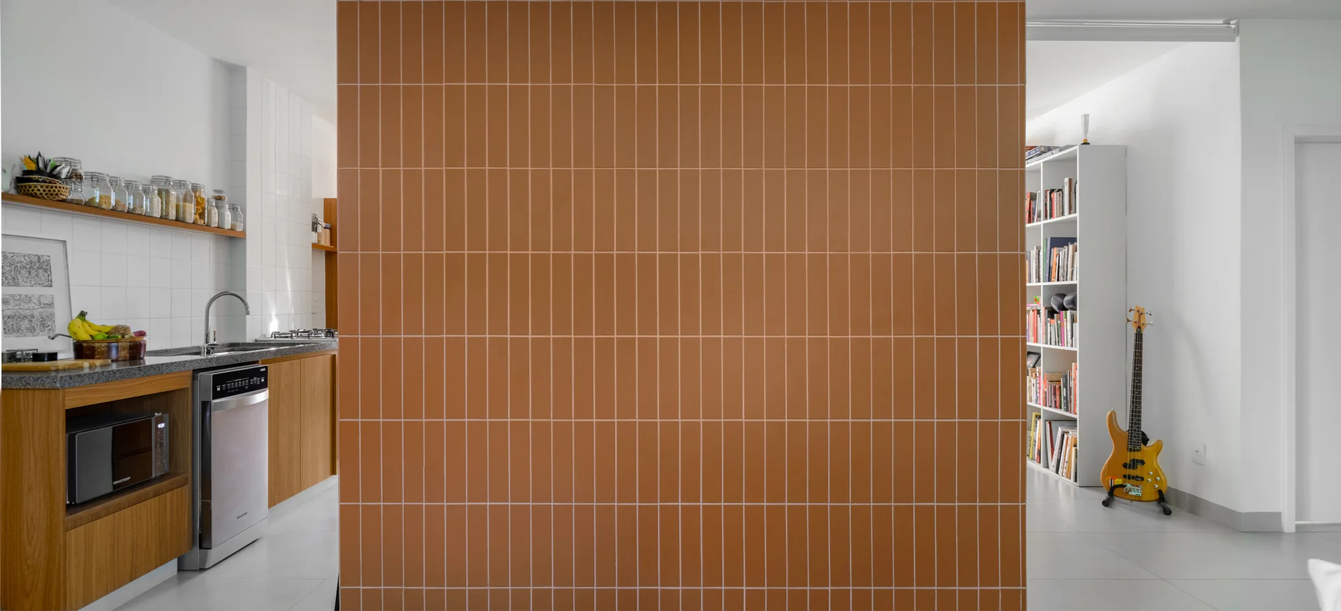A terracotta tiled room divider separates a modern wooden kitchen from a white living area with a bookshelf and bass guitar.