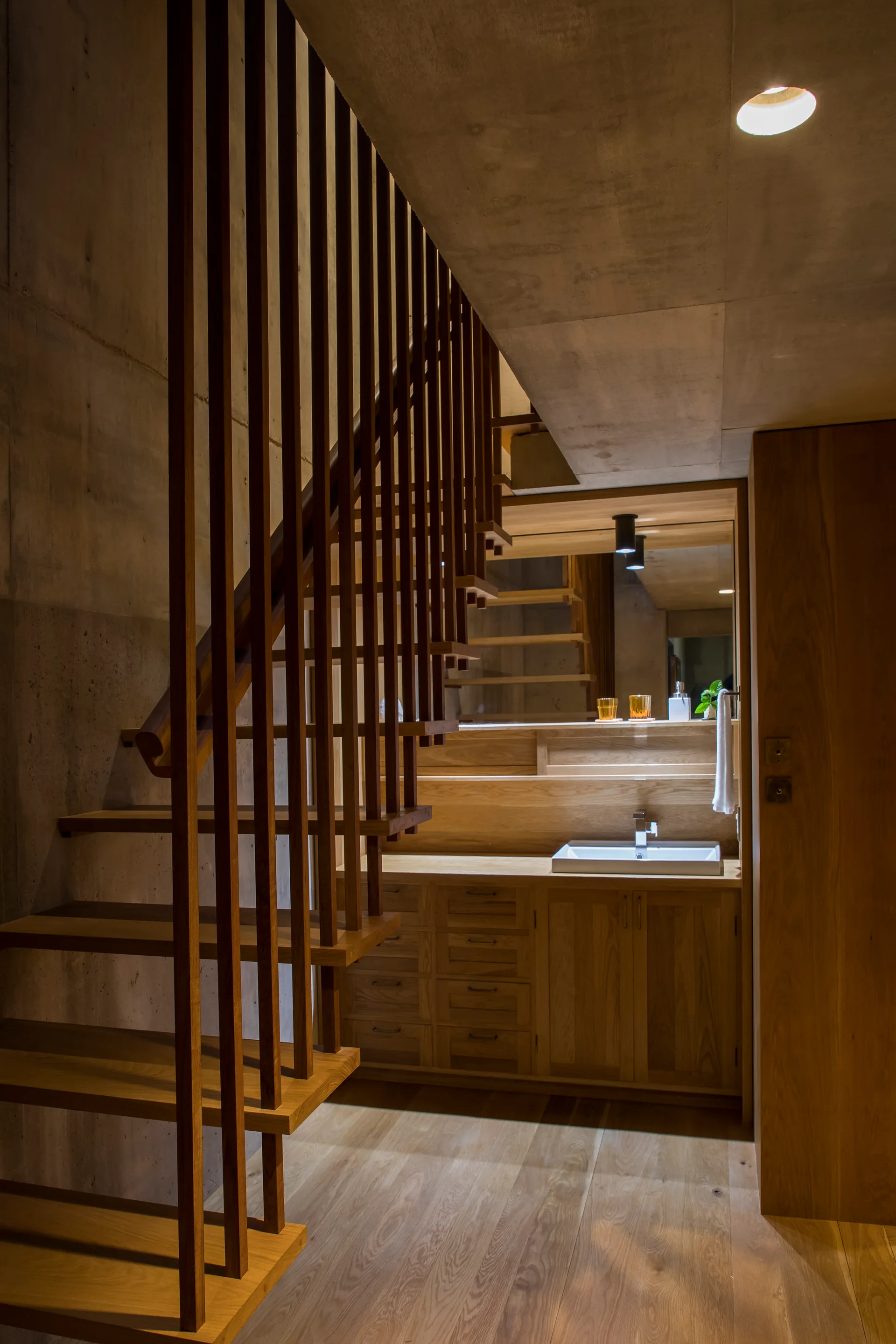 A wooden bathroom vanity with a white sink sits beneath a staircase framed by vertical wooden slats against exposed concrete walls.
