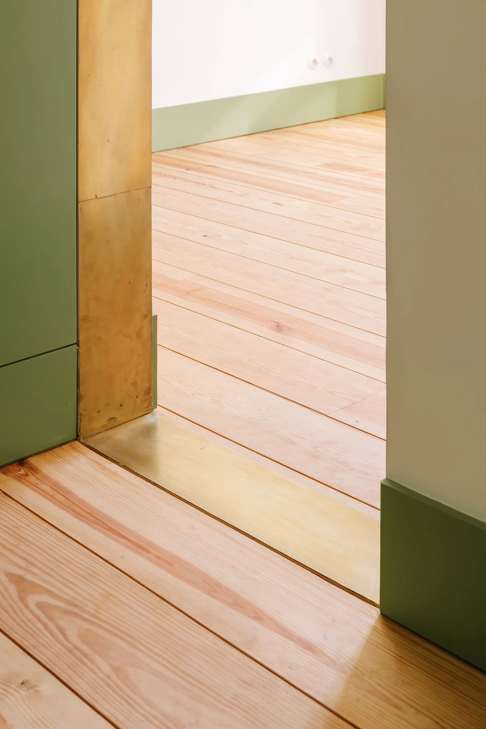 An architectural doorway transition featuring light wood flooring, a polished brass threshold, and sage green painted trim.