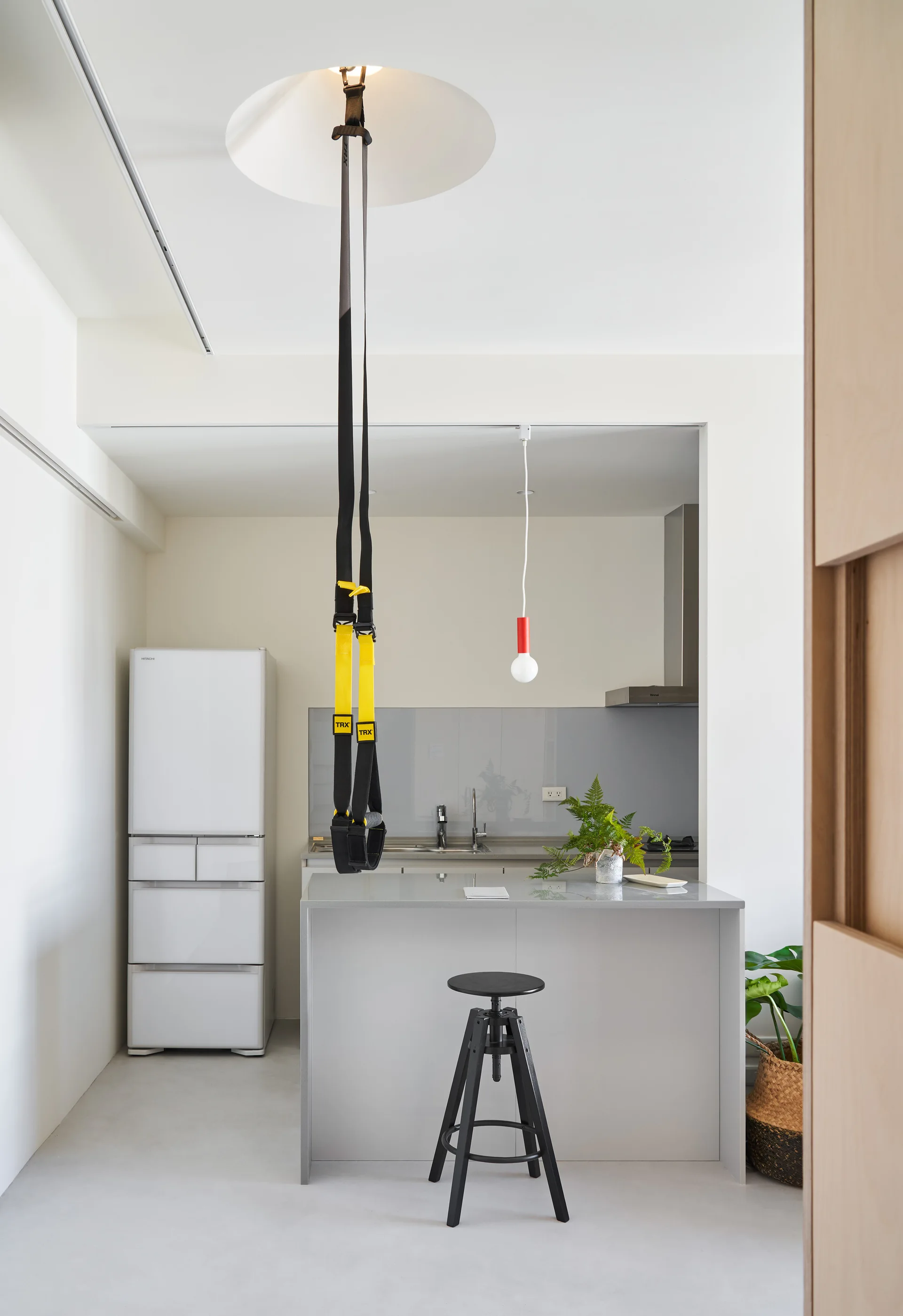 A minimalist kitchen features a gray island with a black stool, a white refrigerator, and black suspension training straps hanging from the ceiling.