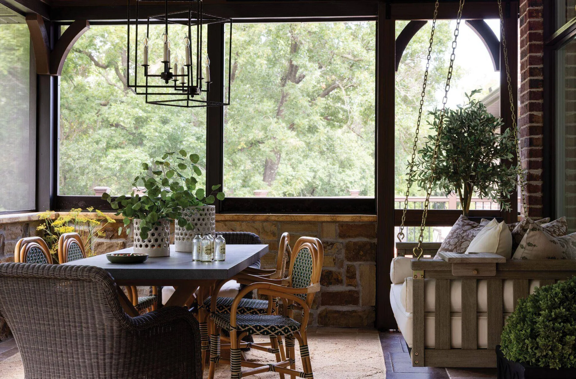 A screened-in porch features a grey dining table with woven rattan chairs alongside a cushioned wooden hanging swing and a stone half-wall.