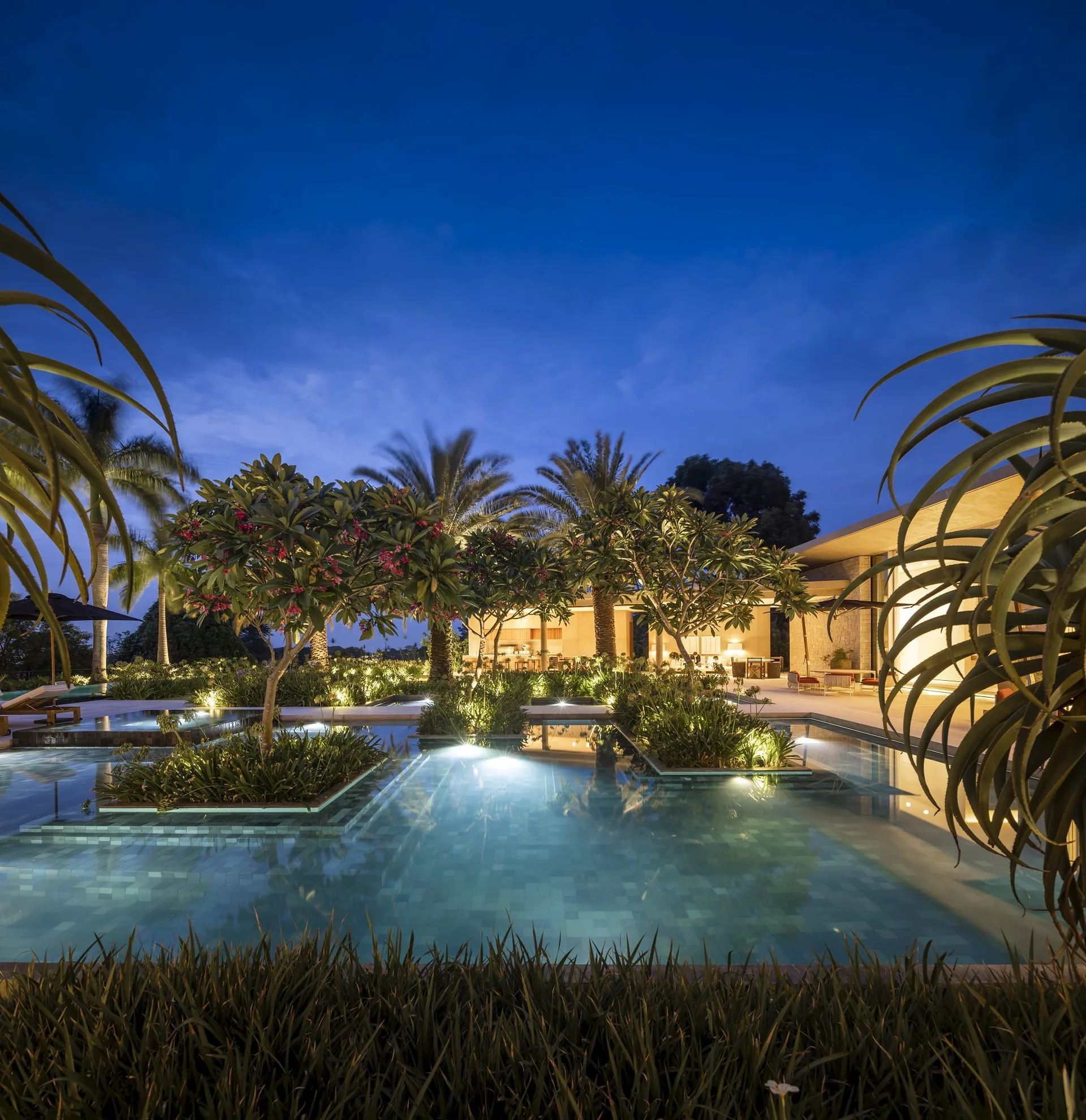 A tiered outdoor swimming pool at dusk surrounded by illuminated tropical plantings and a modern stone structure.