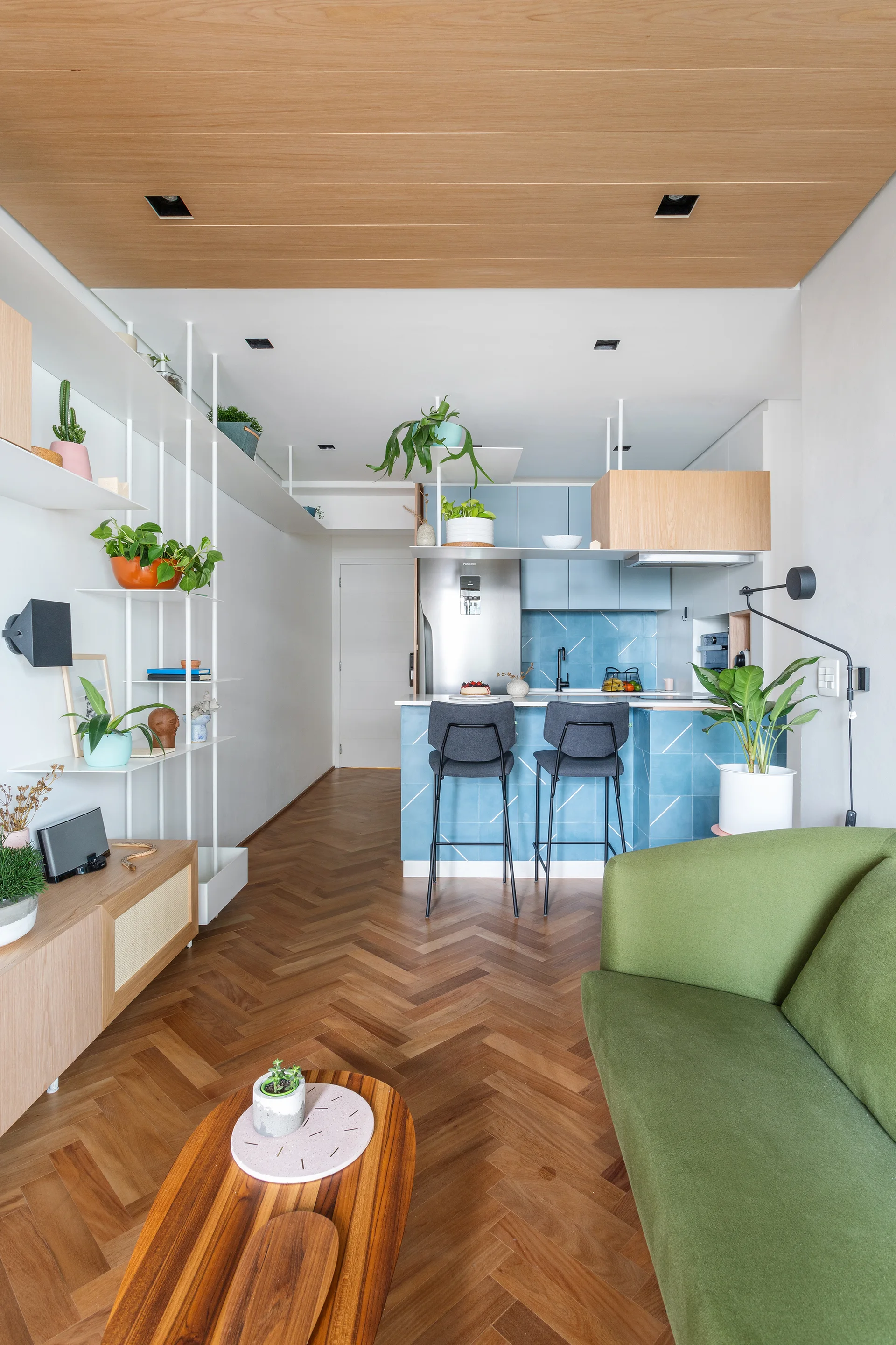 A modern apartment living area features a green sofa, herringbone wood floors, a wood-paneled ceiling, and a kitchen with blue tiles in the background.