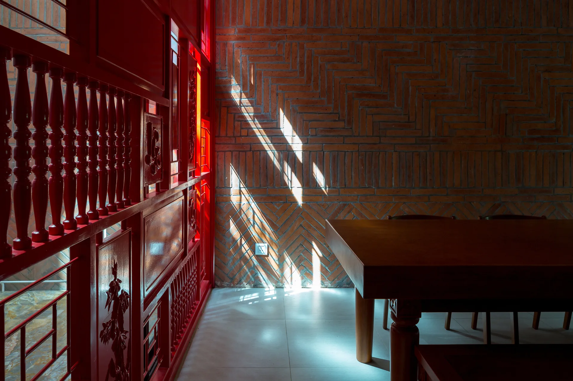 A dining room features a solid wood table sitting before a terracotta herringbone brick wall and a glossy red carved wood partition.