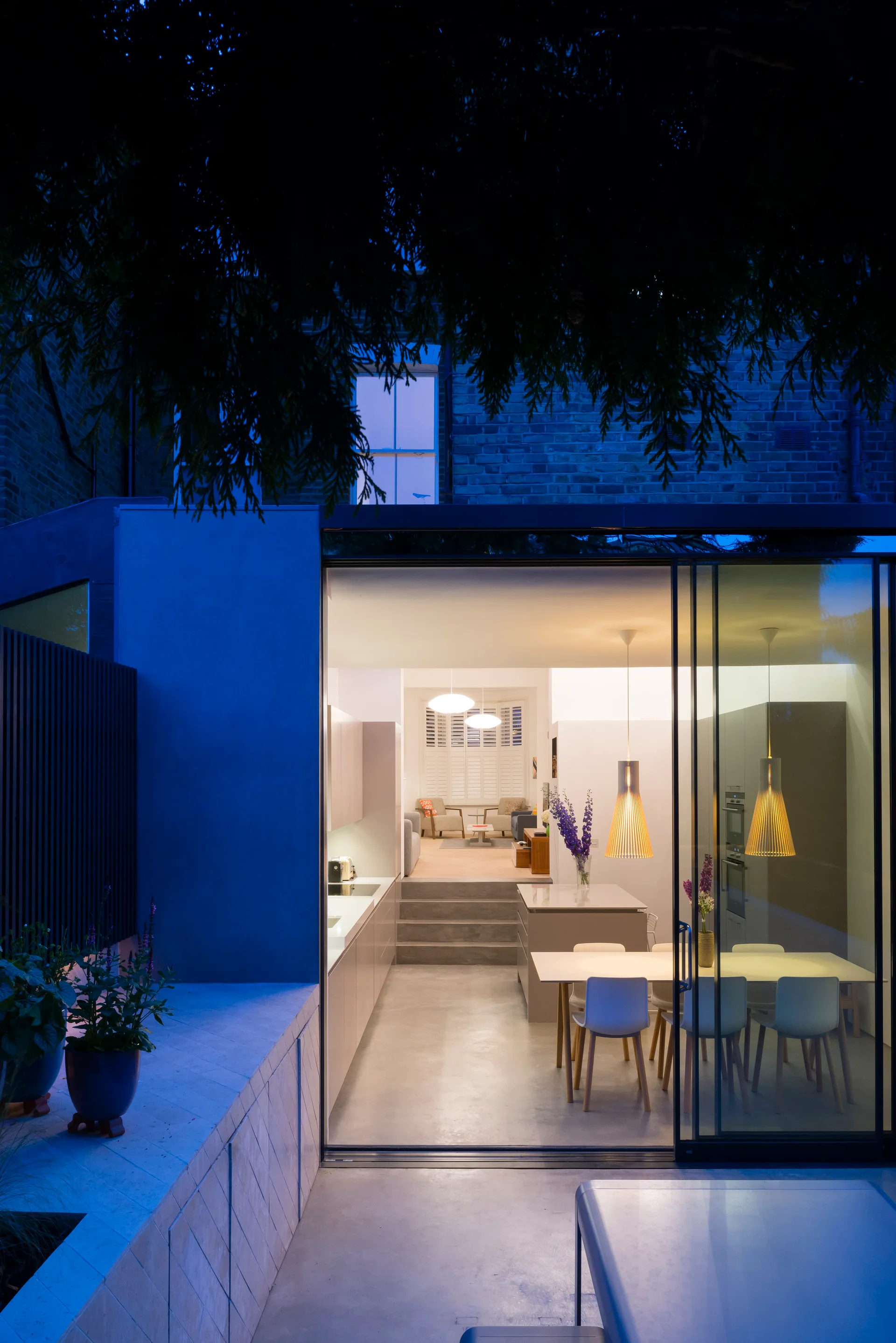 An open-plan kitchen and dining area with a split-level floor and concrete steps, seen through open sliding glass doors from an outdoor patio.
